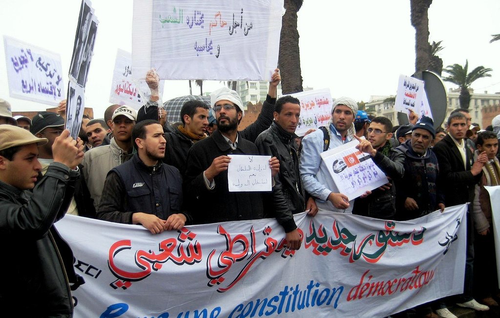 L'image montre un groupe de personnes lors d'une manifestation. Elles tiennent des pancartes et une grande banderole sur laquelle est inscrit en arabe et en français "Pour une constitution démocratique". Les manifestants semblent unis dans leur revendication, et l'environnement montre qu'ils se rassemblent probablement dans une ville, avec des palmiers en arrière-plan. L'atmosphère semble engagée et déterminée.