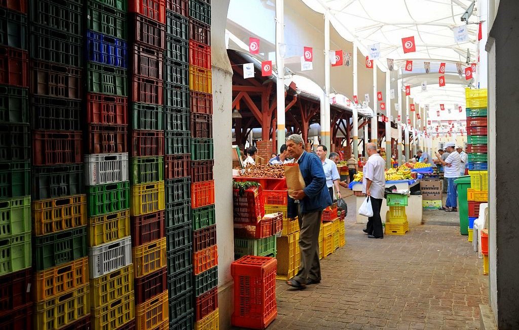 L'image montre un marché couvert animé, avec des étals bien organisés. On peut y voir des rangées de caisses colorées, de différentes tailles et en plastique, empilées de manière ordonnée. Des personnes se déplacent dans le marché, certaines portant des sacs, tandis que d'autres semblent inspecter les produits disponibles. Le tout est sous une structure avec un toit, permettant de se préserver du soleil. Des drapeaux tunisiens décorent le plafond, ajoutant une touche locale à l'ambiance du lieu.
