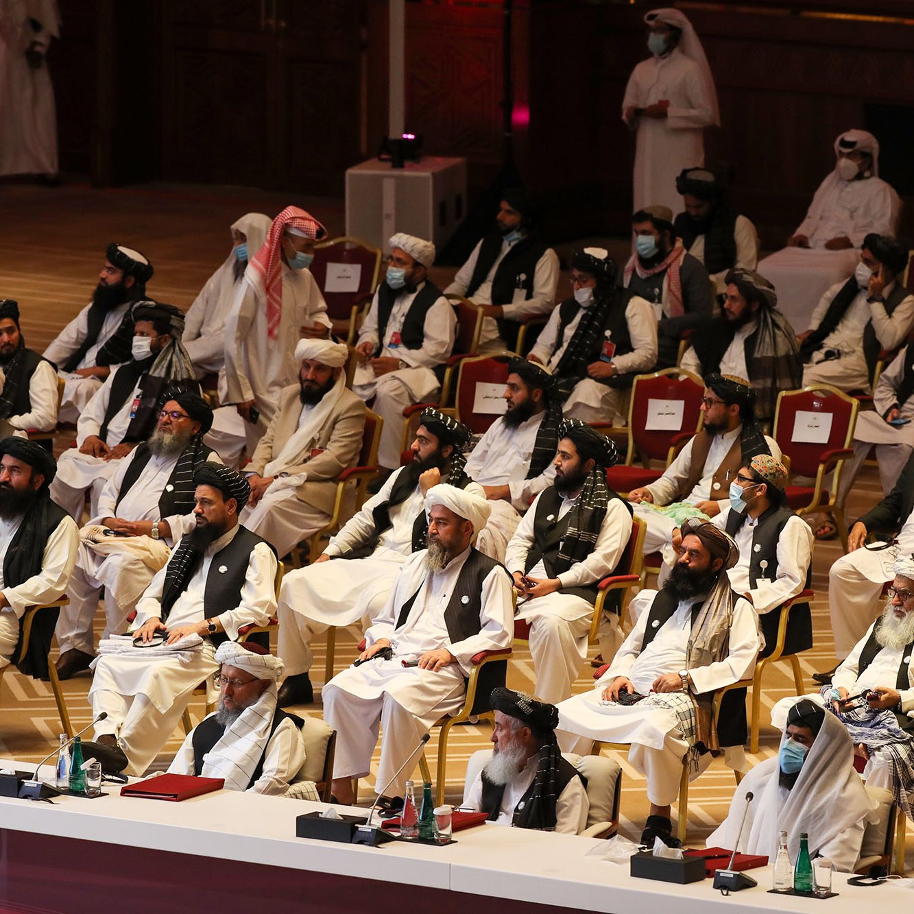 L'image montre un grand rassemblement de personnes assises dans une salle. Les participants sont principalement vêtus de vêtements traditionnels, tels que des turbans et des robes longues. Certains portent des masques. Les gens semblent attentifs et concentrés sur l'événement en cours. L'environnement est formel, avec des sièges disposés en rangées, et une scène à l'avant. L'atmosphère générale évoque une réunion officielle ou un congrès.