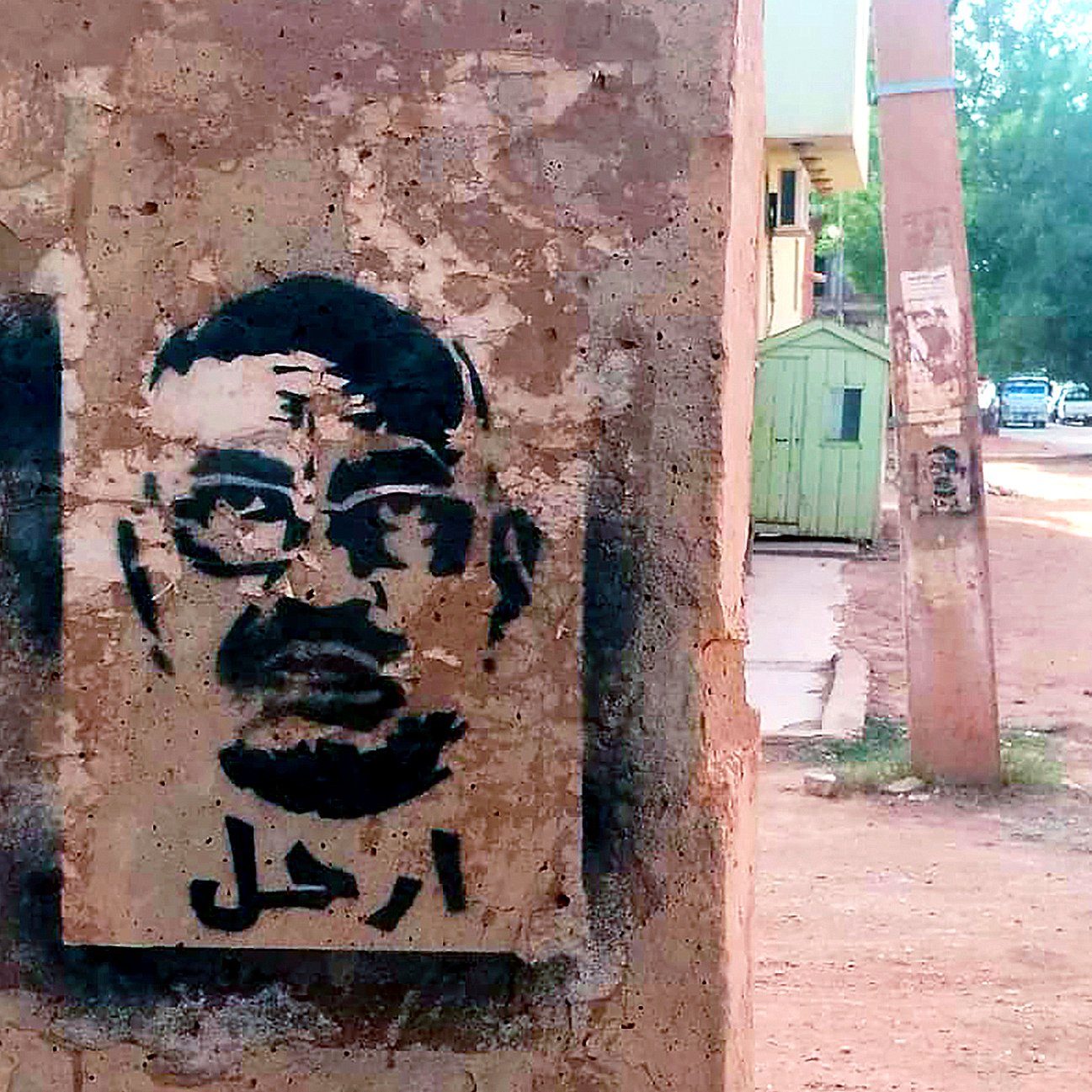 L'image montre un graffiti sur un mur, représentant un visage stylisé d'un homme avec des lunettes. Le dessin est en noir, sur un fond de béton clair. En arabe, le mot "ارحل" est inscrit, ce qui signifie "pars" ou "sors". L'environnement semble urbain, avec des bâtiments et une route visible en arrière-plan.