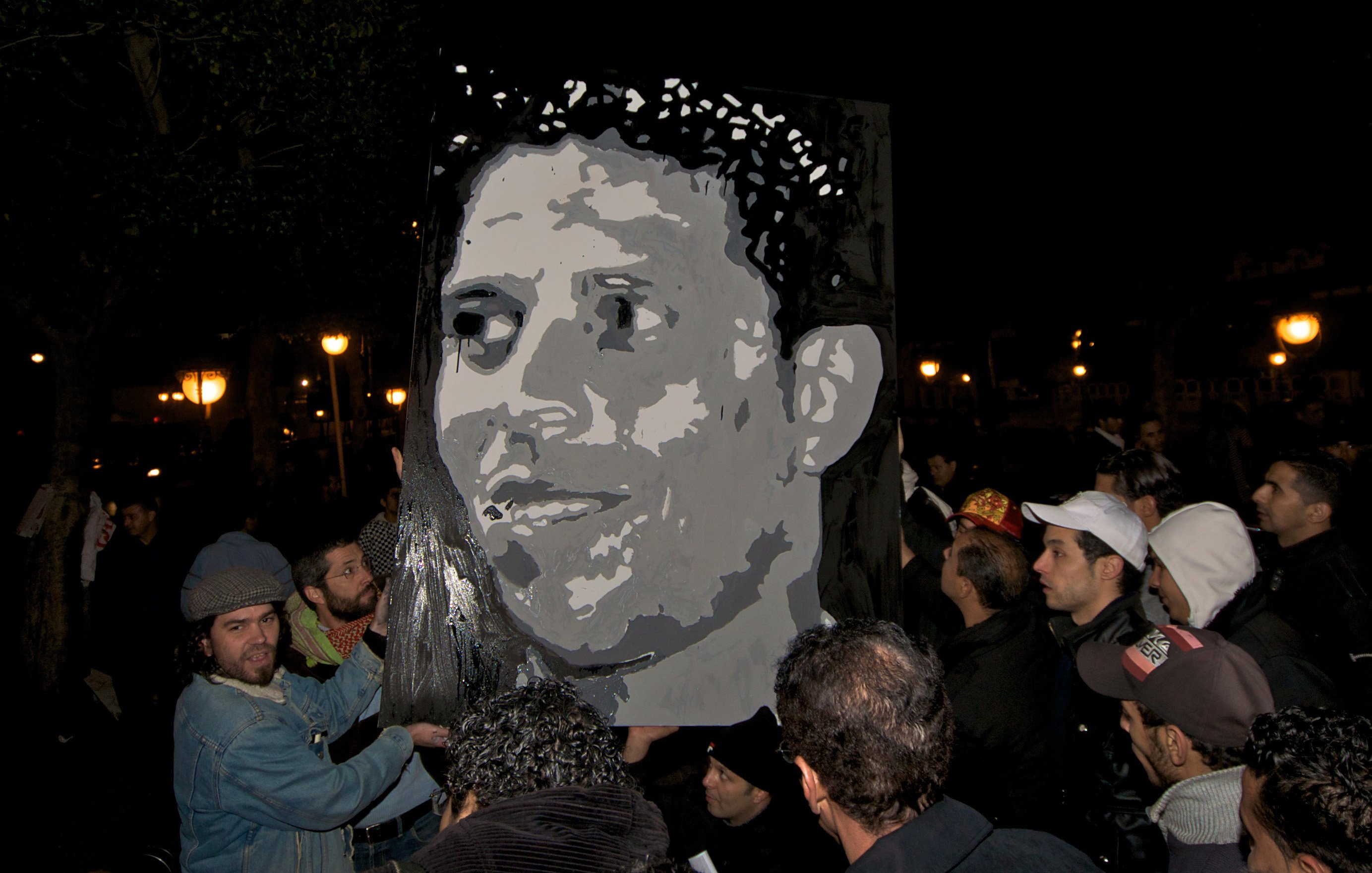 L'image montre un groupe de personnes rassemblées dans un environnement nocturne. Au centre, une grande affiche ou un portrait d'un homme est visible, avec des couleurs sombres et une expression marquante. Les personnes semblent engagées et intéressées par le portrait qu'elles tiennent, ce qui suggère qu'il s'agit d'une manifestation ou d'un hommage. L'ambiance nocturne est accentuée par l'éclairage des lampadaires en arrière-plan.