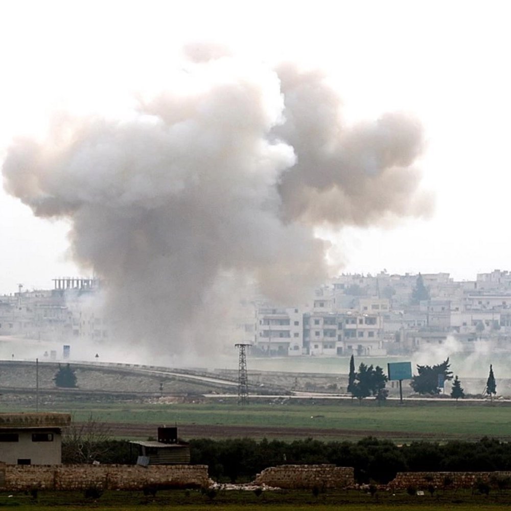 تصویر یک صحنه جنگی را نشان می‌دهد که در آن دود و گرد و غبار ناشی از یک انفجار دیده می‌شود. در پس‌زمینه، ساختمان‌های شهری و یک منطقه باز مشاهده می‌شود. آسمان ابری است و حالت کلی تصویر به موقعیتی پر تنش و خطرناک اشاره دارد. دود در حال بلند شدن از زمین به سمت آسمان است و نشانه‌هایی از ویرانی و نبرد در تصویر مشهود است.