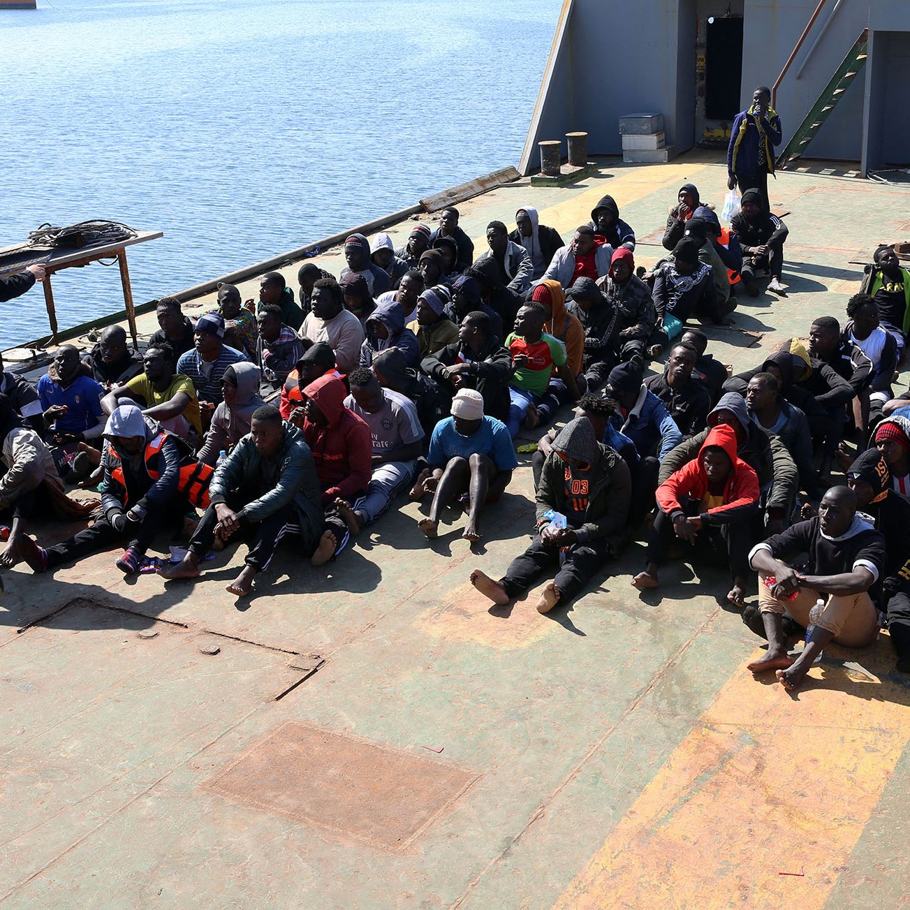 L'image montre un groupe de personnes assises sur le pont d'un bateau. Elles semblent être des migrants, plusieurs portant des vêtements chauds et des masques. À l'arrière, une personne en uniforme militaire observe la scène, tandis qu'un autre individu est vêtu d'un gilet portant un logo d'une organisation humanitaire. L'environnement semble être une zone portuaire, avec de l'eau visible à l'arrière-plan. L'atmosphère de l'image évoque une situation de contrôle ou d'assistance humanitaire.