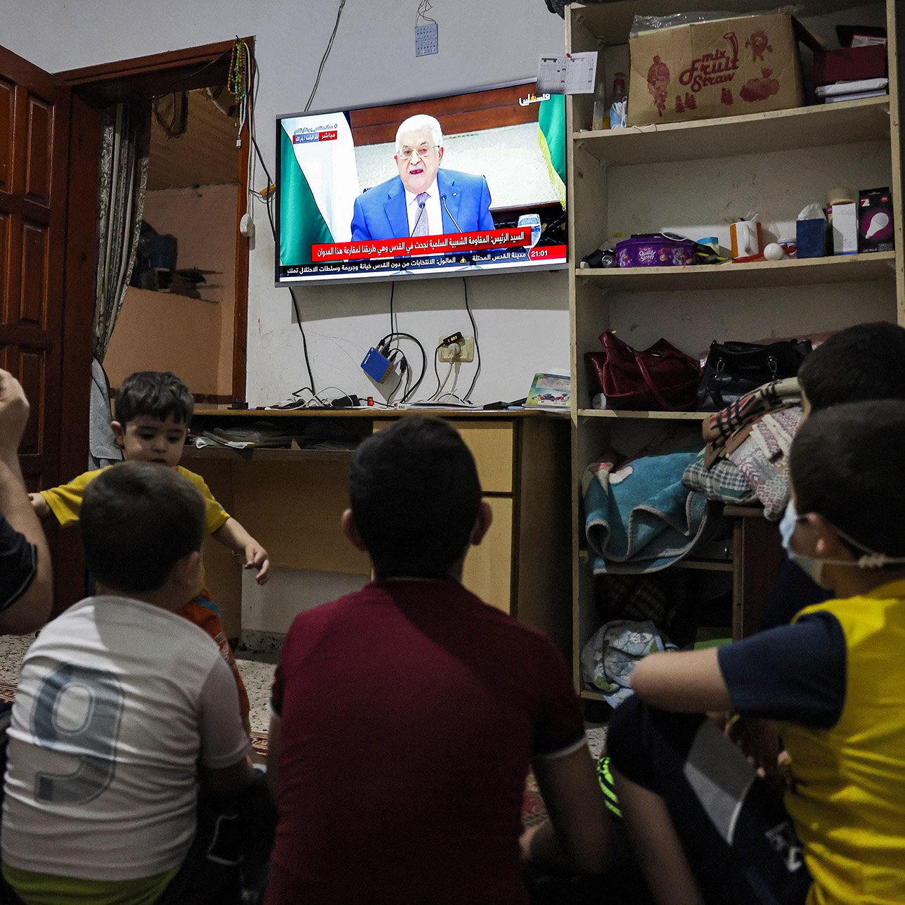 L'image montre un groupe d'enfants assis sur le sol, regardant la télévision. Un adulte, probablement un parent, est également présent et semble interagir avec les enfants. La télévision affiche un homme parlant, probablement un discours ou une intervention publique. L'environnement est simple, avec des meubles basiques et des objets éparpillés. Les enfants semblent absorbés par ce qu'ils voient à l'écran.
