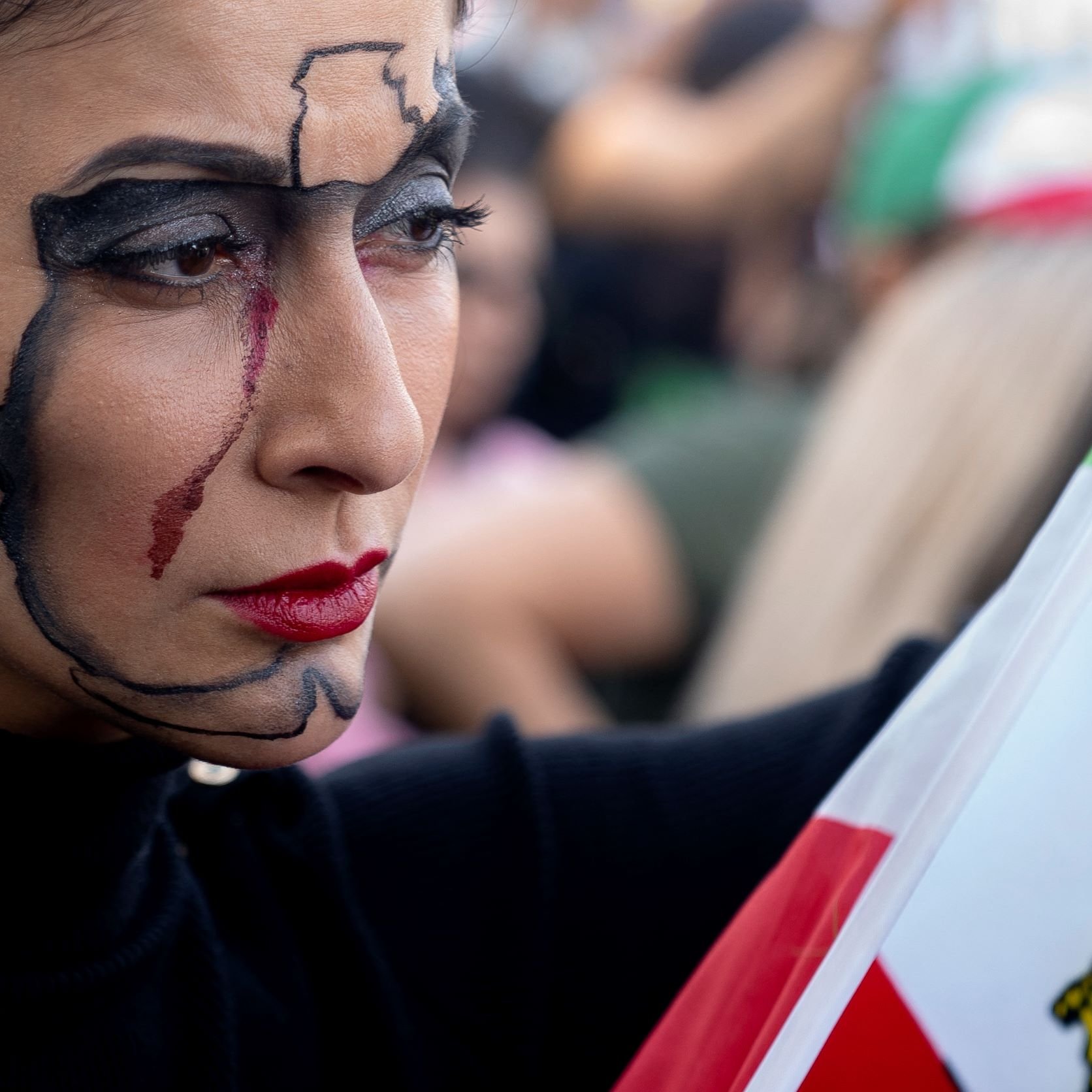 L'image montre une femme avec un maquillage artistique qui représente une carte. Elle a des lignes noires lisses sur son visage qui dessinent une forme géographique, accompagnées d'une larme rouge. Sa bouche est accentuée par un rouge à lèvres également rouge. Elle tient un drapeau, probablement national, avec des couleurs distinctives. L'arrière-plan semble flou, avec d'autres personnes en mouvement, suggérant un rassemblement ou une manifestation.