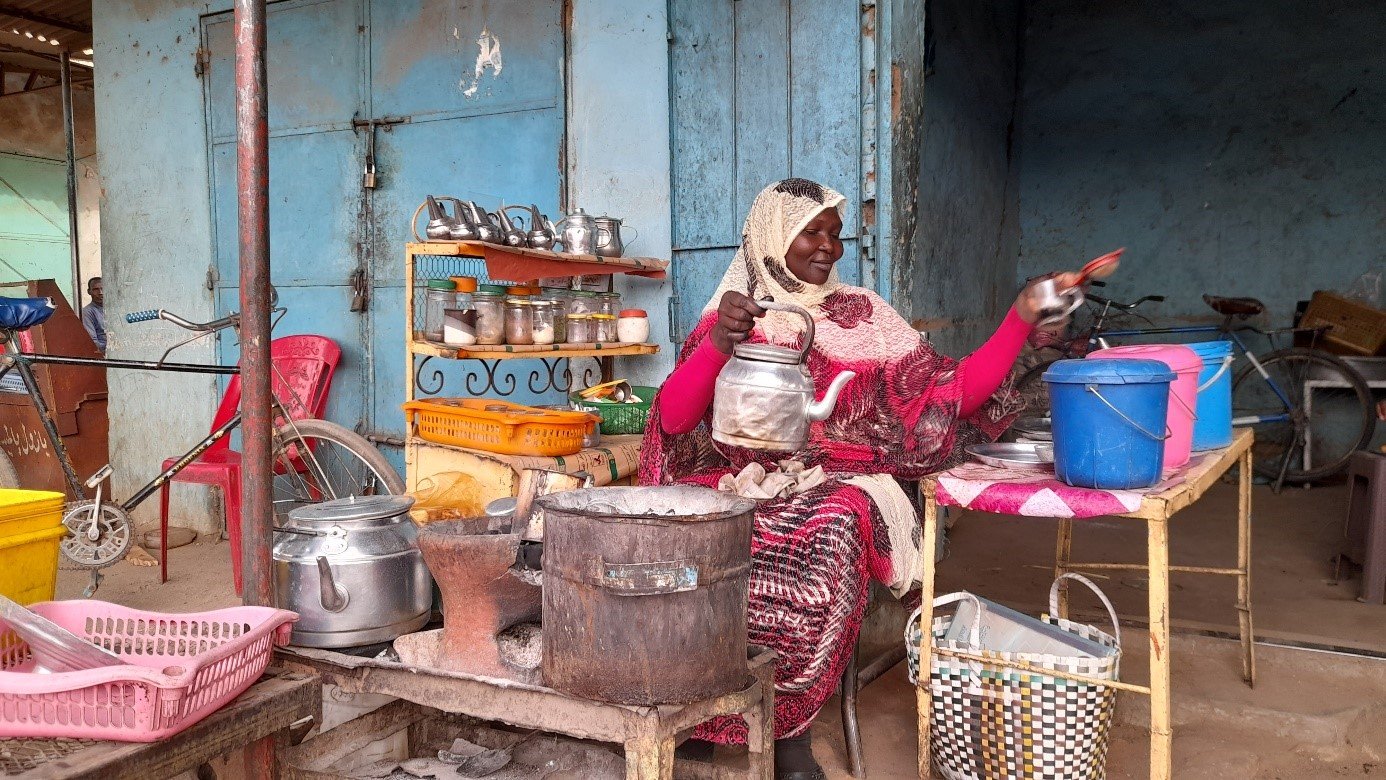 L'image montre une femme assise devant un petit stand ou un kiosque. Elle porte un vêtement traditionnel, probablement coloré, et a un foulard sur la tête. Autour d'elle, on peut voir divers ustensiles de cuisine et des récipients, comme des casseroles et des seaux en plastique de différentes couleurs. Sa table est en métal et semble utilisée pour préparer ou vendre de la nourriture. En arrière-plan, on aperçoit des murs peints et d'autres objets, créant une atmosphère vivante et locale.