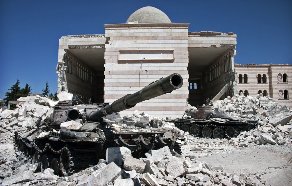 L'image montre un paysage de destruction, probablement lié à un conflit. Au premier plan, on voit des chars endommagés, couverts de débris et de poussière. En arrière-plan, il y a un bâtiment partiellement détruit, dont les murs présentent des marques des destructions. Le ciel est bleu, ce qui contraste avec la scène de désolation au sol. L'ensemble évoque un endroit marqué par la guerre, avec des traces visibles de violence et de désastre.