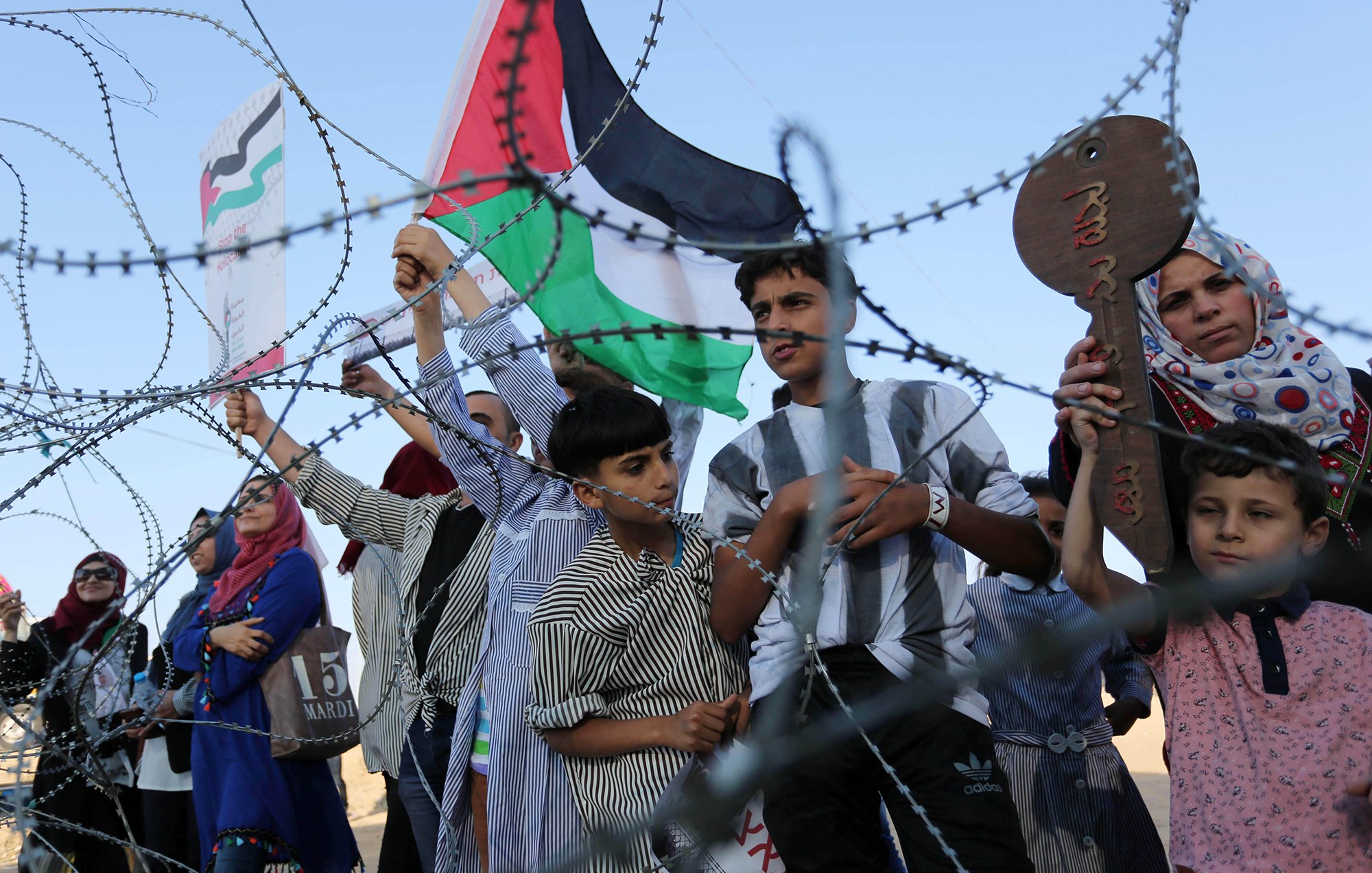 L'image montre un groupe de personnes, y compris des enfants, qui tiennent des drapeaux palestiniens et des pancartes. Ils se tiennent derrière une barrière semblable à du barbelé. Les expressions des personnes peuvent communiquer des émotions telles que la détermination ou l'espoir. L'environnement semble être en plein air, sans autre détail contextuel visible.