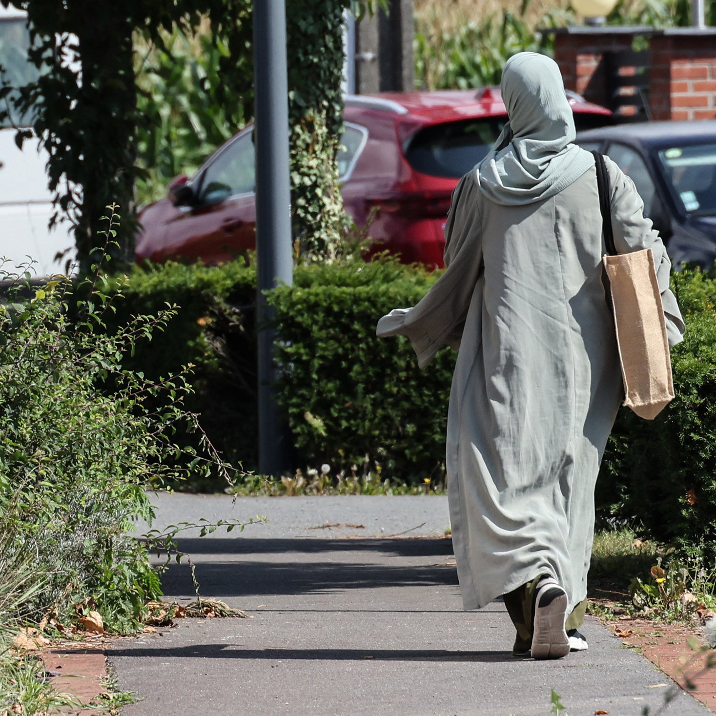 L'image montre une personne marchant sur un chemin. Elle porte une longue robe de couleur claire et un foulard. Elle tient un grand sac en papier dans une main. Le décor environnant inclut des buissons et des arbres, avec des voitures en arrière-plan. La scène semble tranquille et ensoleillée.