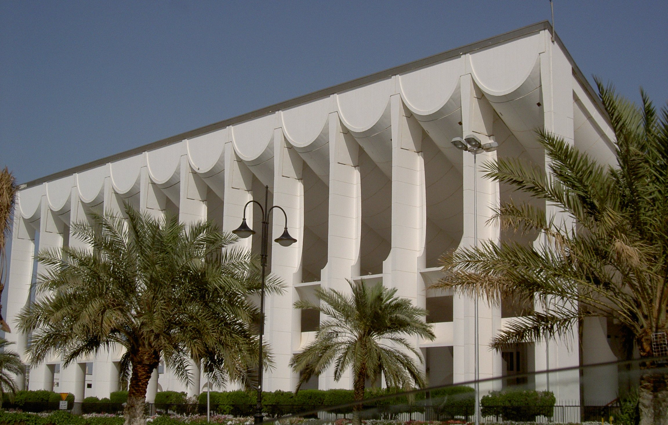 L'image montre un bâtiment moderne avec une architecture distinctive. La façade est principalement blanche, ornée de structures en forme de vagues ou de voiles sur le haut. Des colonnes soutiennent la structure, ajoutant une touche de sophistication. On peut également observer des palmiers plantés devant le bâtiment, ce qui donne une atmosphère tropicale et accueillante. Le ciel est clair et bleu, accentuant la luminosité de l'ensemble.