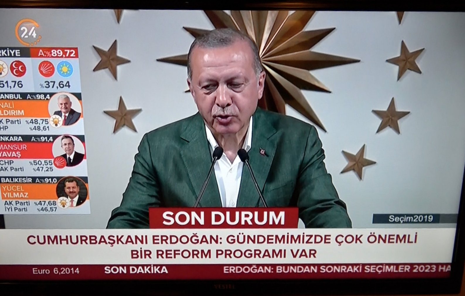 L'image montre un homme en train de prendre la parole lors d'une conférence, probablement un discours politique. Il porte une veste verte et se tient devant un grand fond décoré, avec ce qui semble être un symbole national ou institutionnel. En bas de l'écran, il y a un bandeau rouge qui indique "SON DURUM" et mentionne "CUMHURBAŞKANI ERDOĞAN" avec un message sur des réformes importantes. Sur le côté gauche, des informations sur des élections et des résultats sont affichées, suggérant que l'événement est lié à des élections en Turquie.