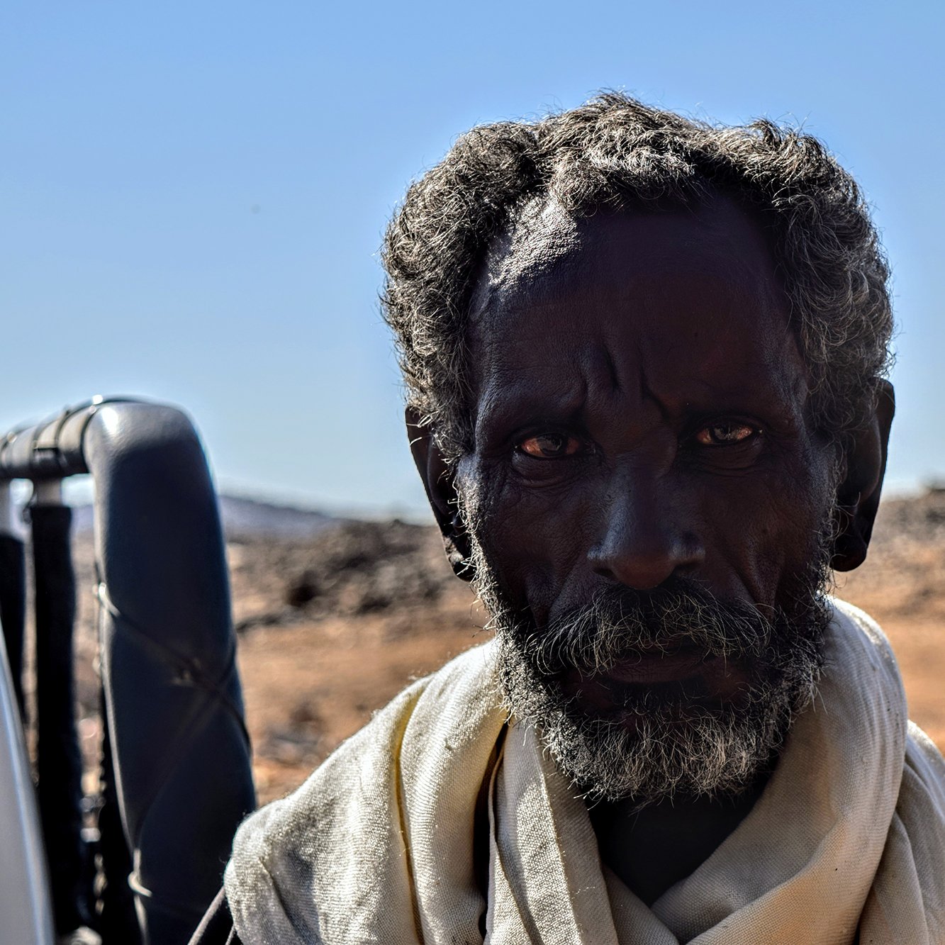 الصورة تظهر رجلاً مسنًا ذا لحية وشعر مجعد، يبدو عليه علامات الزمن والخبرة. عينيه تعكسان نظرة واضحة وقوية، بينما يتواجد في خلفية طبيعة جافة تتضمن صخور وتضاريس غير مستوية. يظهر الرجل بملابس بسيطة، مما يعكس بساطة الحياة في المنطقة التي يعيش فيها. السماء زرقاء والجو يبدو صافياً.