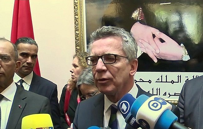 تُظهر الصورة شخصًا يتحدث أمام مجموعة من الكاميرات والميكروفونات، ويبدو أنه في مؤتمر صحفي أو حدث رسمي. خلفه، يوجد صورة كبيرة ليد مع بعض الرموز، ربما تتعلق بالتراث أو الثقافة. هناك أيضًا عدد من الأشخاص الآخرين يقفون في الخلفية، مما يشير إلى أهمية الحدث أو اللقاء. الأعلام التي تظهر في الصورة تعزز الطابع الرسمي للموقف.