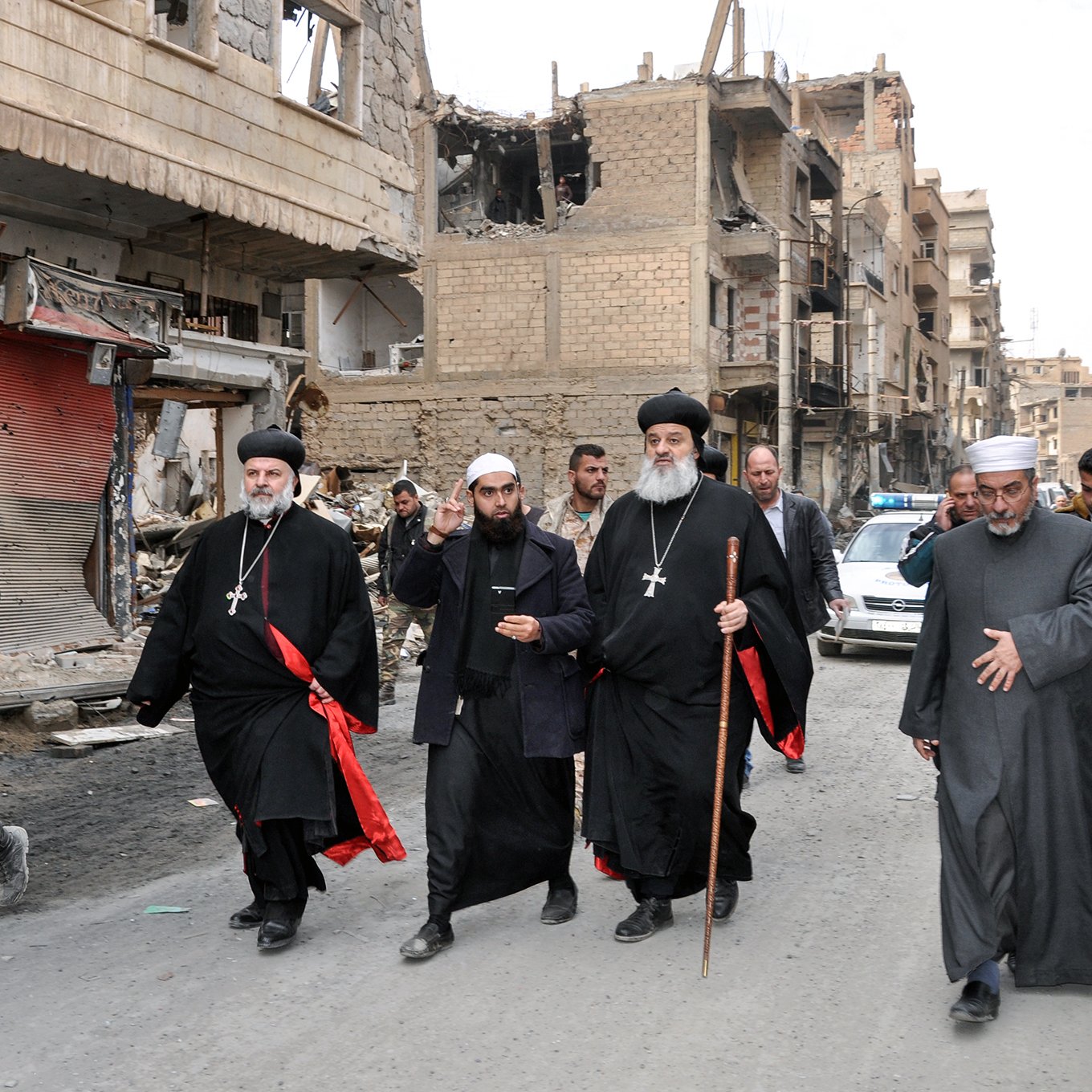 The image shows a group of religious figures walking through a war-torn street. They are dressed in traditional clergy attire, including black robes and crosses, and some are holding staffs. The surroundings are heavily damaged, with buildings showing signs of destruction and debris on the street. In the background, there are a few people and vehicles, indicating a somber atmosphere in a once-bustling area. The overall scene reflects the impact of conflict on the community and highlights the presence of religious leadership amidst the destruction.