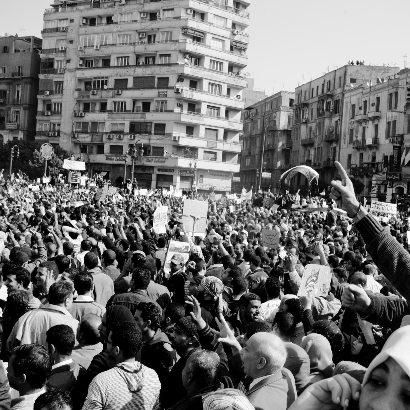 L'image montre une grande foule rassemblée dans une ville, probablement lors d'une manifestation ou d'une protestation. Les participants brandissent des pancartes et semblent exprimer des opinions fortes. L'atmosphère est dynamique et engagée, avec beaucoup de personnes levées les bras, ce qui suggère une ambiance de mobilisation collective. Le noir et blanc de l'image accentue les émotions et l'intensité du moment. Les bâtiments environnants ajoutent un contexte urbain à cette scène de rassemblement.