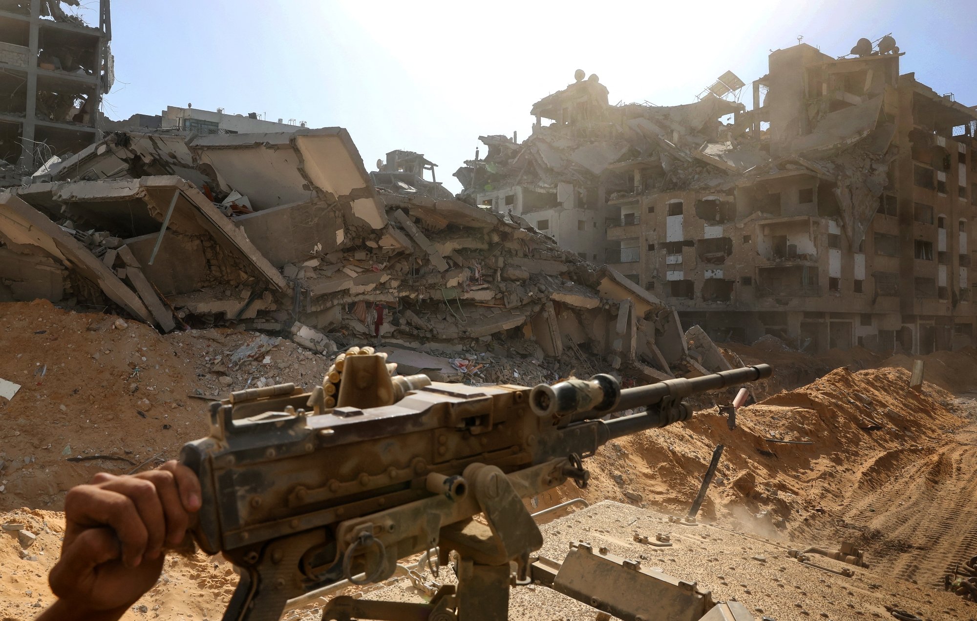 تصویری از یک منطقه ویران‌شده، با ساختمان‌های خراب و تفنگی در پیش‌زمینه. غبار و تابش نور خورشید در آسمان.