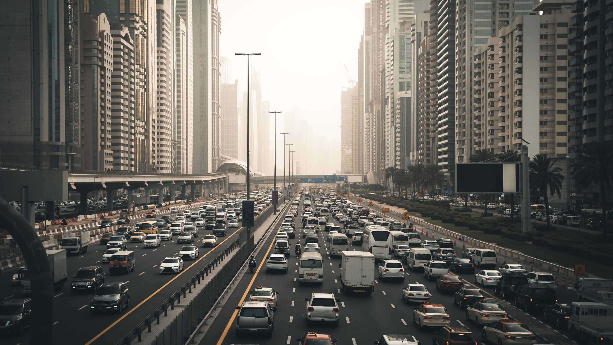 تظهر في الصورة مشهدًا حضريًا لمدينة مزدحمة، حيث تتواجد الكثير من السيارات المتوقفة في زحام مروري على عدة مسارات. تحيط بالمكان ناطحات السحاب العالية التي تدل على التطور العمراني في المنطقة. تبدو السماء مغشاة بضباب خفيف، مما يعطي انطباعًا عن أجواء المدينة في وقت متأخر من اليوم أو خلال الصباح الباكر. هناك مساحات من الأشجار على جوانب الطريق تضفي لمسة من الطبيعة وسط الحضر.
