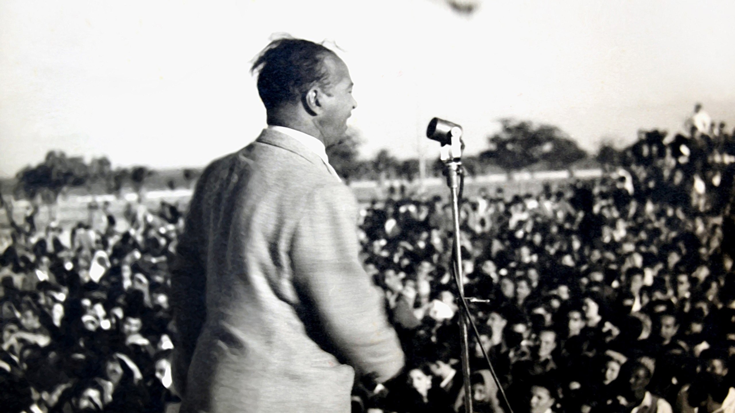 L'image montre un homme en train de parler sur une scène, devant un grand public. Il est vu de dos, et on peut voir une foule nombreuse devant lui, captivée par son discours. On distingue également un microphone sur le podium. L'atmosphère semble dynamique et engagée, probablement lors d'un événement politique ou d'un rassemblement. En arrière-plan, il y a des drapeaux, ce qui pourrait indiquer un contexte symbolique ou patriotique.