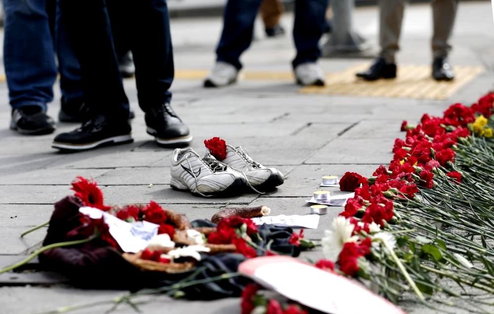L'image montre un sol avec une paire de chaussures de sport blanches laissées à l'écart. Autour des chaussures, il y a une traînée de fleurs rouges, probablement des œillets, qui sont disposées en ligne. On peut également voir d'autres objets sur le sol, tels que des photos et des éléments décoratifs. En arrière-plan, des gens sont visibles, mais leur apparence et leurs détails ne sont pas clairement discernables. L'ensemble de la scène dégage une atmosphère de mémoire ou de commémoration.