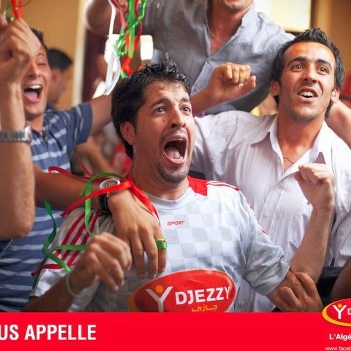 L'image montre un groupe de jeunes hommes dans une ambiance festive. Ils semblent très joyeux et enthousiastes, certains criant et souriant, probablement en train de célébrer un événement sportif. Ils portent des vêtements décontractés et il y a des décorations colorées autour d'eux. Au premier plan, un homme exprime sa joie de manière très expressive, avec les bras levés. En bas, on peut voir un slogan qui dit "LA VIE VOUS APPELLE". L'ensemble de la scène dégage une atmosphère de camaraderie et de bonheur collectif.