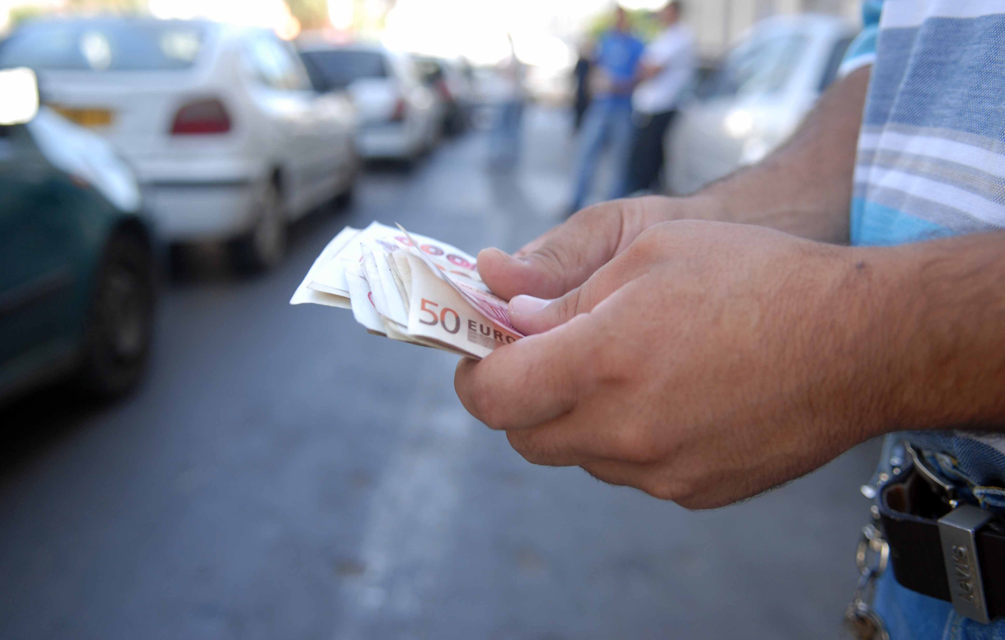 L'image montre des mains tenant des billets de 50 euros. En arrière-plan, on peut voir des voitures garées le long d'une rue. L'ambiance semble être celle d'un environnement urbain. Les mains sont fermement posées sur les billets, ce qui suggère une transaction ou le comptage d'argent.