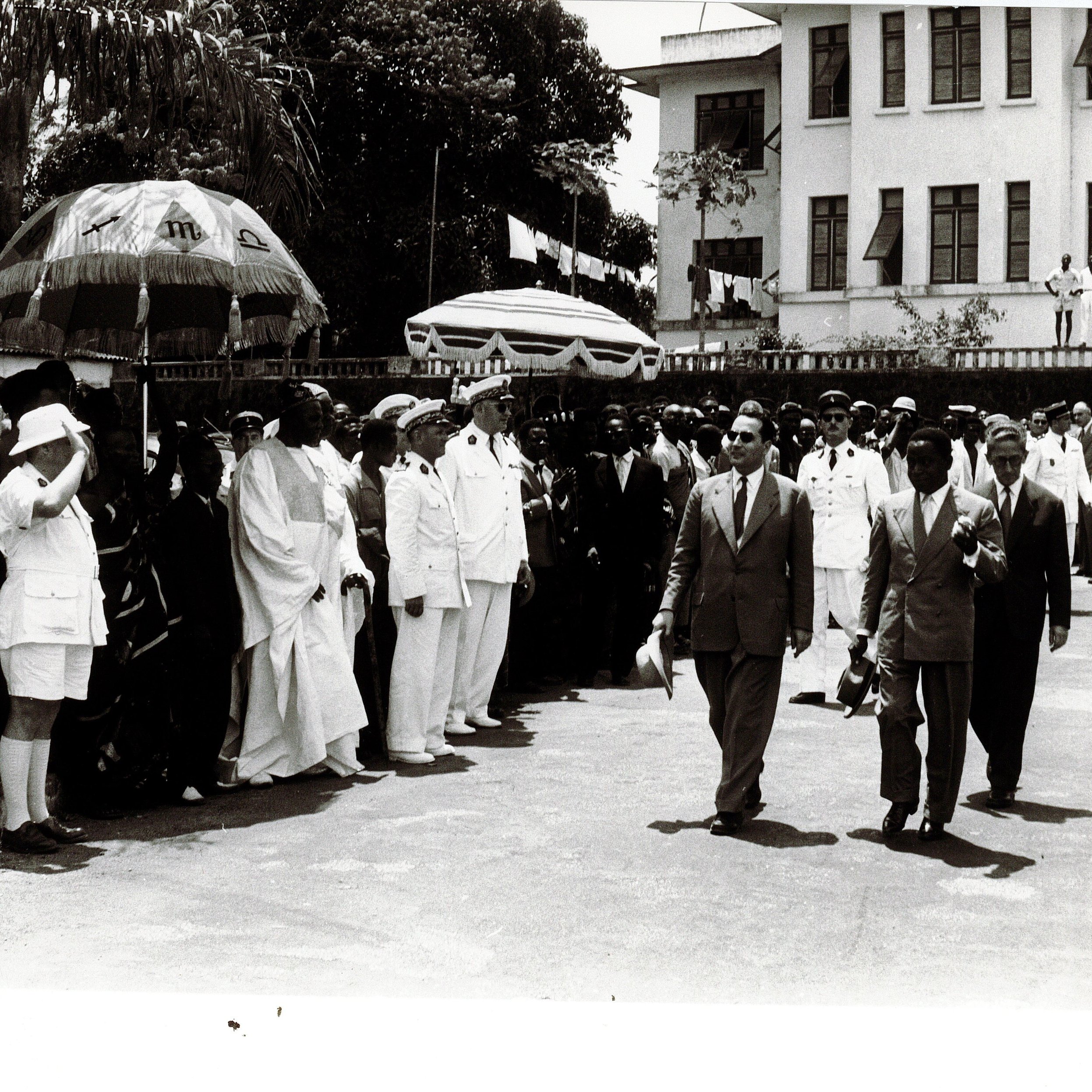 L'image montre une scène publique, probablement une cérémonie officielle. On y voit un groupe de personnes en costume, regroupées sur un côté, avec des hommes portant des chapeaux et des vêtements formels. À l'avant, on distingue des hommes en uniformes militaires et d'autres en tenue élégante. Au centre, une figure principale semble marcher vers la caméra, entourée de gardes et d'accompagnateurs. Des parasols colorés sont également visibles, ajoutant une touche d'ombre à la scène. En arrière-plan, on peut apercevoir un bâtiment officiel avec des drapeaux, indiquant un événement important ou une célébration. L'atmosphère semble solennelle et organisée.
