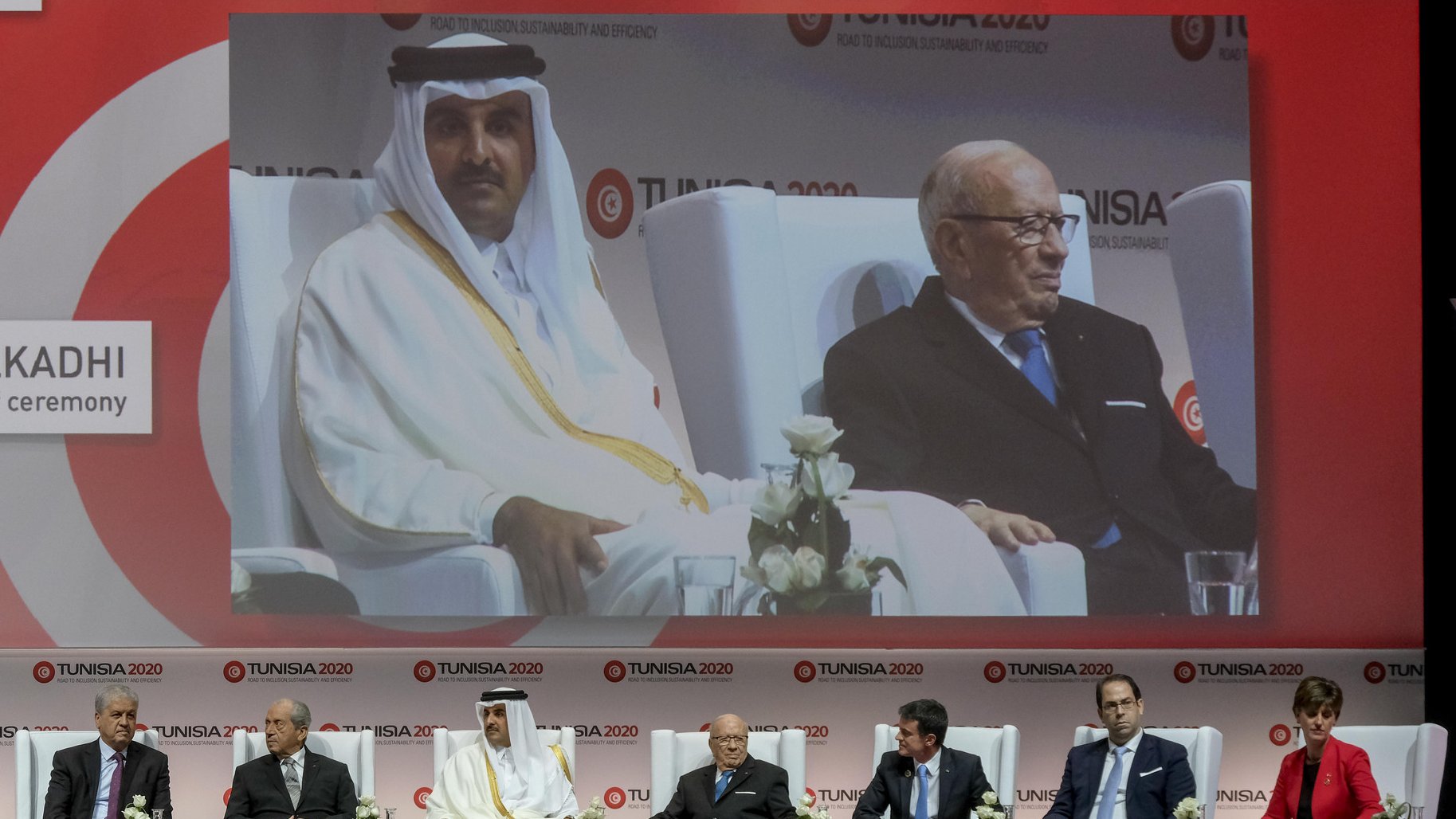 L'image montre une scène de conférence ou de séminaire avec plusieurs intervenants assis sur des chaises, sur une scène décorée avec des fleurs blanches. Au centre, il y a un grand écran affichant un homme en costume, vraisemblablement un orateur ou un participant à l'événement. Les participants semblent représentés sur une plateforme, prenant part à une discussion ou à une cérémonie. Le fond est décoré avec des motifs et des logos liés à l'événement "TUNISIA 2020". L'ambiance semble professionnelle et formelle.