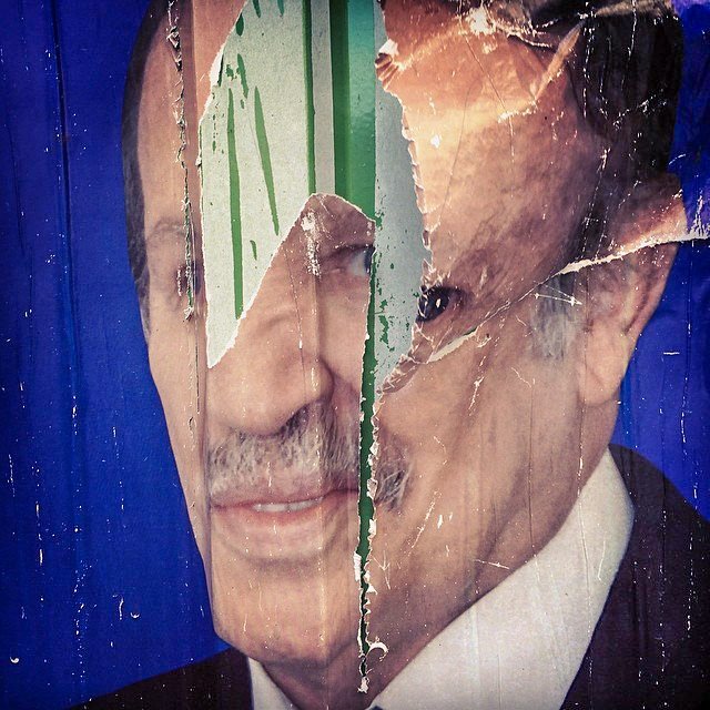 L'image montre un affichage déchiré d'un visage, probablement d'une personne publique, sur un fond bleu. Le visage est partiellement caché par des déchirures et des marques de pluie ou de peinture verte, ce qui donne un aspect usé et dégradé à l'image. Les détails du visage, comme les yeux et la bouche, sont visibles en partie, mais l'ensemble est affecté par le temps et l'usure.