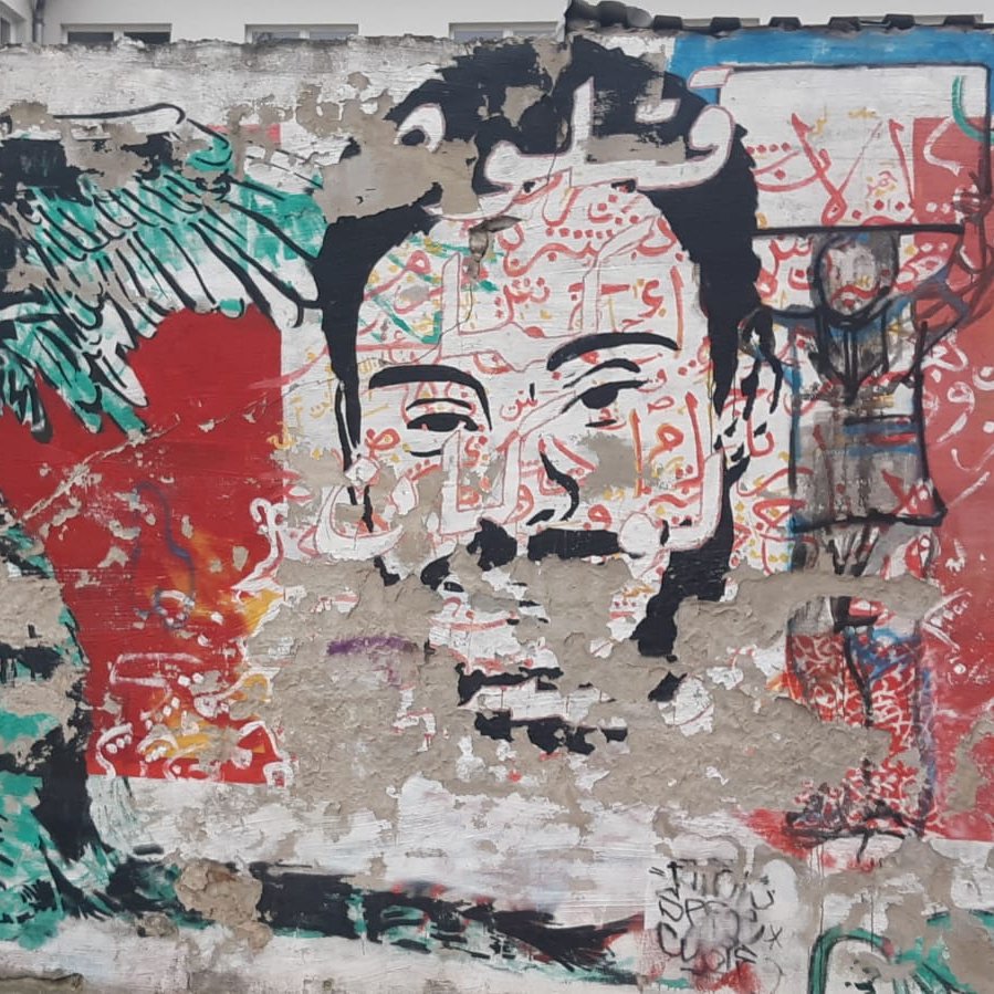 L'image montre une fresque murale riche en couleurs et en détails. À gauche, on peut voir un personnage avec des ailes et un visage expressif, qui semble être une représentation stylisée d'une créature. À droite, se trouve un visage humain qui attire l'attention, entouré de motifs vibrants. En arrière-plan, divers éléments graphiques et des inscriptions en arabe ajoutent une dimension culturelle. L'ensemble donne une impression de dynamisme et de créativité, typique de l'art urbain.