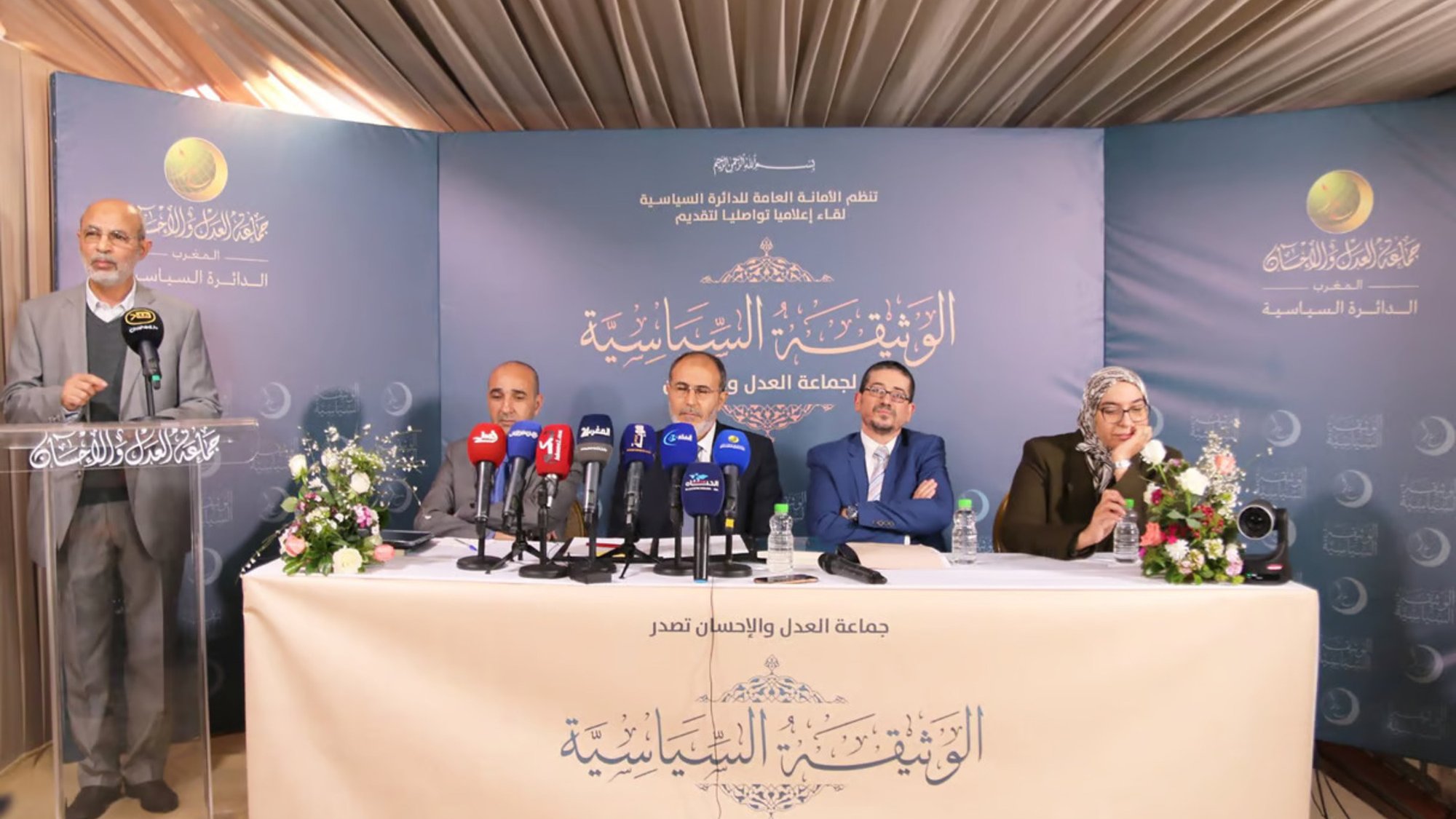 L'image montre une conférence de presse avec plusieurs intervenants assis derrière un pupitre. Ils sont entourés de micros et de caméras, indiquant une attention médiatique. Des fleurs et une décoration soignée ajoutent une touche de professionnalisme à l'événement. On peut voir un intervenant debout, probablement en train de parler ou de présenter, tandis que les autres écoutent attentivement. Le fond de la scène est orné de motifs ou de logos, suggérant que c'est un événement officiel ou formel.