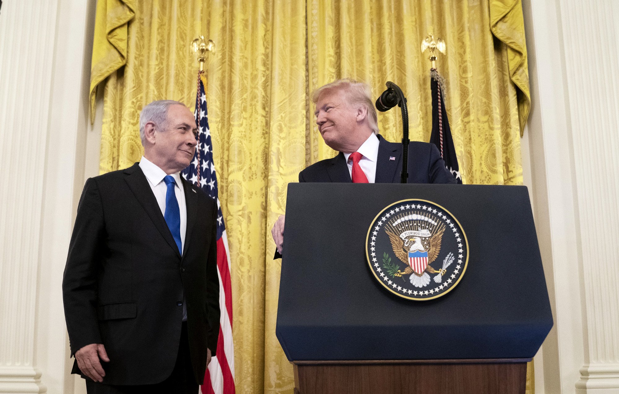 تصویر مربوط به یک مراسم رسمی است که در آن دو نفر در حال گفتگو هستند. در پس‌زمینه، پرده‌های طلایی و پرچم‌های ایالات متحده دیده می‌شوند. یکی از افراد در سمت چپ، لباسی تیره به تن دارد و به شخص دیگر در سمت راست نگاه می‌کند. شخص در سمت راست یک لباس رسمی و یک کراوات به تن دارد و به نظر می‌رسد در حال لبخند زدن است. همچنین بر روی میزی که در مقابل آنها قرار دارد، نشان رسمی ریاست‌جمهوری ایالات متحده وجود دارد. این تصویر به نظر می‌رسد به یک رویداد سیاسی یا دیپلماتیک مربوط باشد.