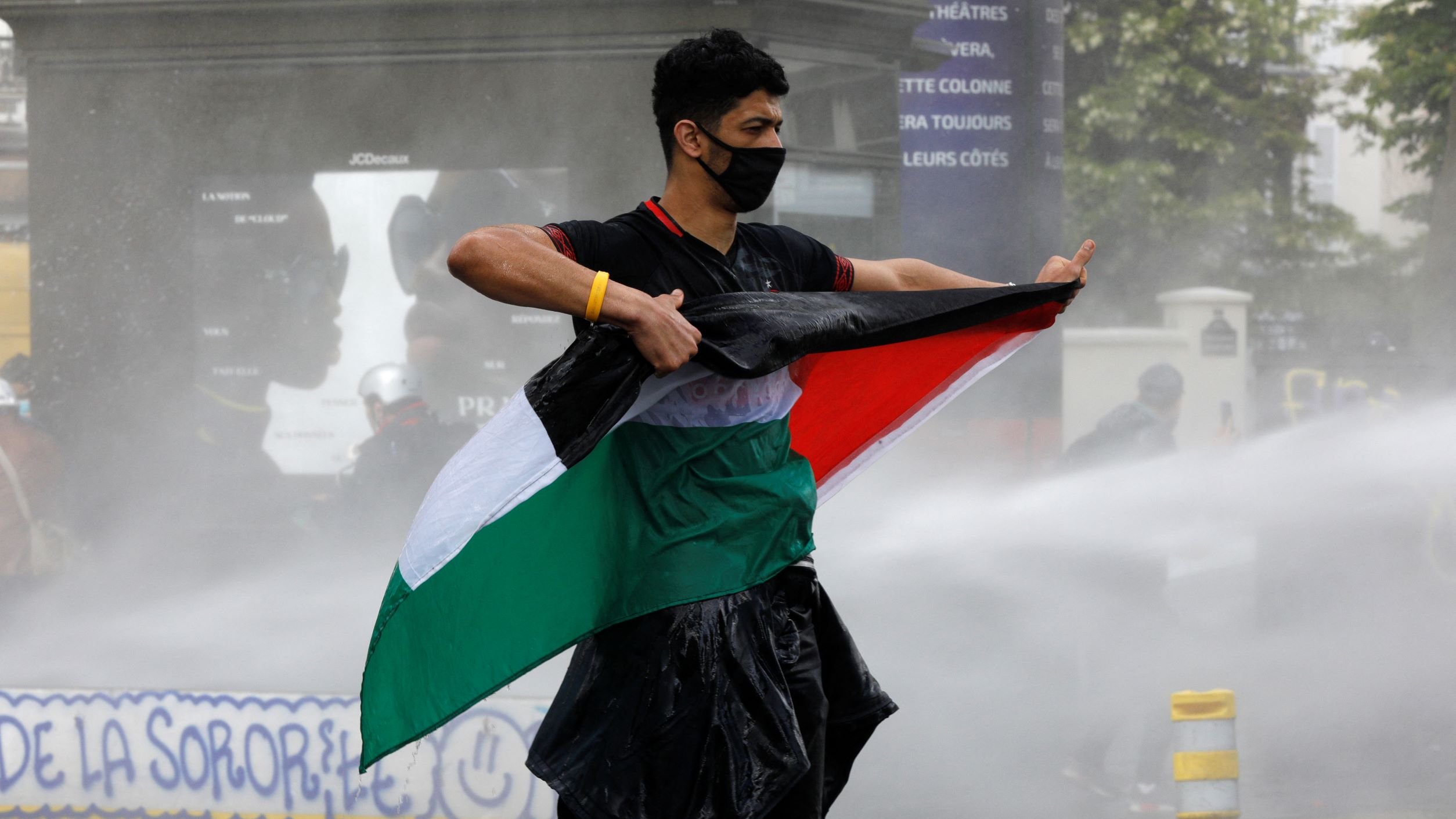 Sur l'image, on voit un jeune homme debout, tenant un drapeau. Il porte un masque noir et semble faire face à une situation de tension, peut-être une manifestation. Le drapeau qu'il brandit a des couleurs verde, blanc et rouge, tandis que de l'eau jaillit autour de lui, suggérant l'utilisation de canons à eau. L'environnement semble être urbain, avec des signes de protestation en arrière-plan.