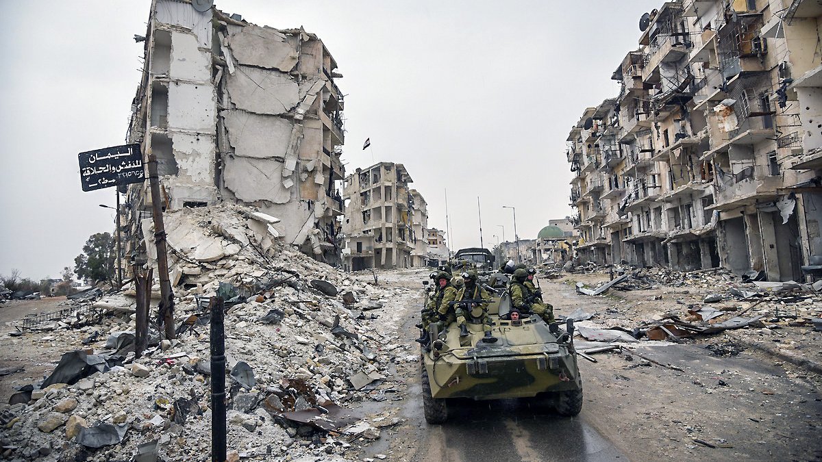 L'image montre une scène de destruction urbaine, probablement à la suite d'un conflit. On peut voir des bâtiments en ruine, avec des façades effondrées et des débris éparpillés sur le sol. Au premier plan, il y a un véhicule militaire, sur lequel des soldats sont présents. L'atmosphère semble sombre et chargée, soulignant les ravages causés par la guerre.