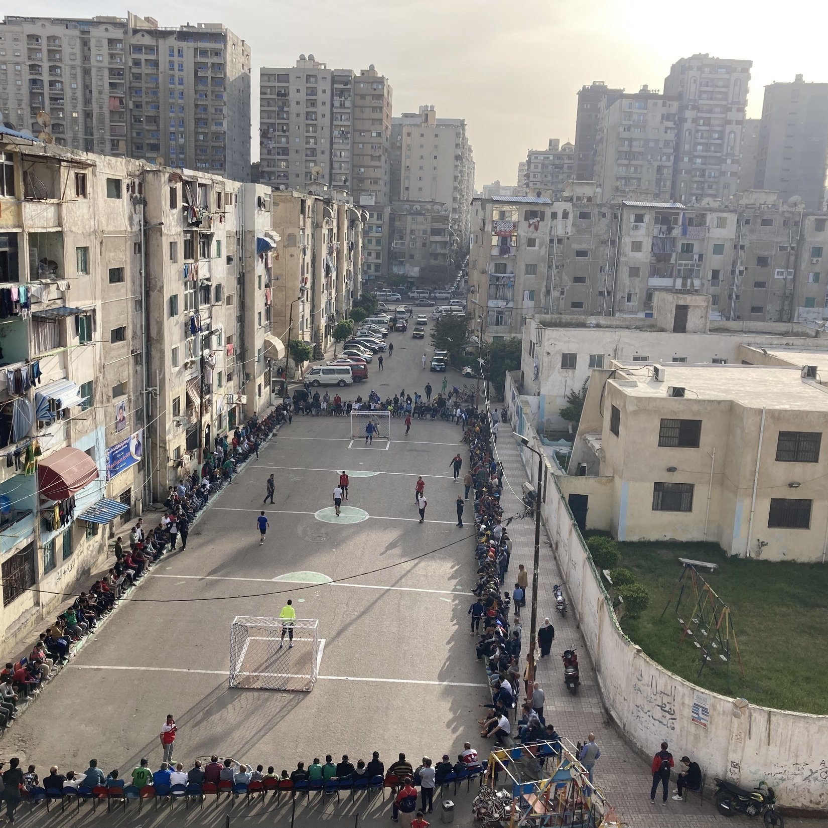 L'image montre un terrain de football encadré par des immeubles d'apartements dans une zone urbaine. Plusieurs personnes sont rassemblées sur des bancs le long du terrain, probablement en train de regarder un match. On peut voir des joueurs en action sur le terrain. L'établissement est entouré de bâtiments de plusieurs étages, témoignant d'une ambiance animée et communautaire. L'éclairage du soir ajoute une atmosphère chaleureuse à la scène.