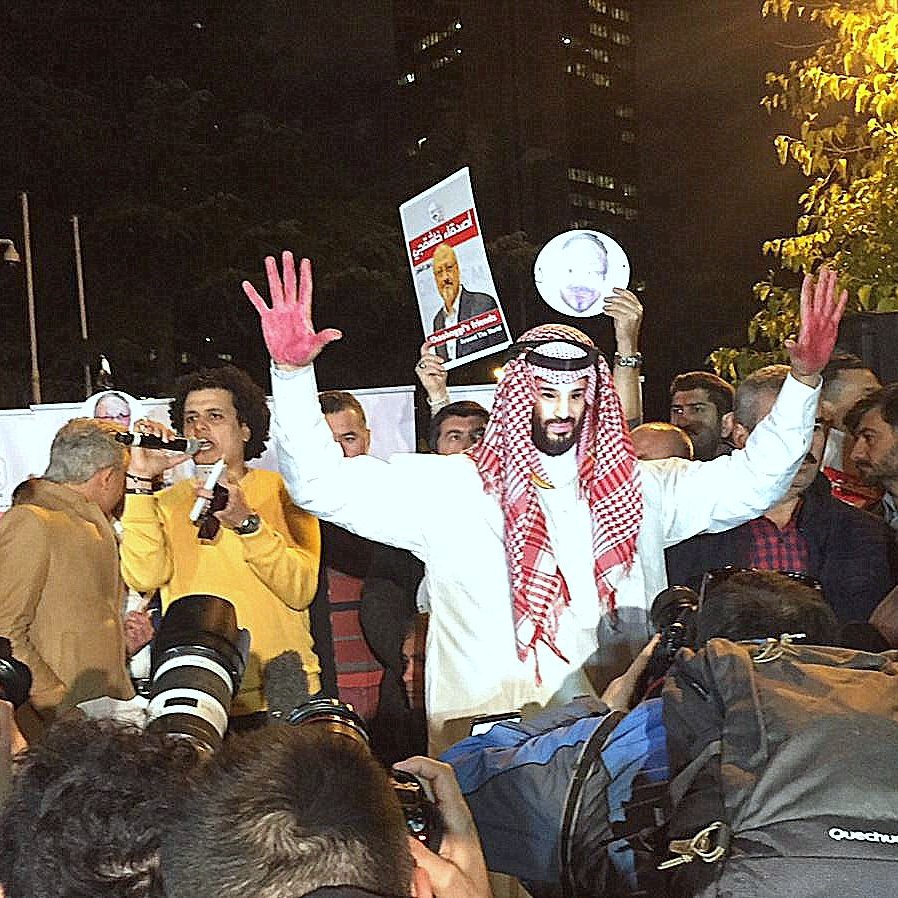 تظهر الصورة مجموعة من الأشخاص في حدث عام أو احتجاج. في الوسط، يوجد رجل يرتدي ملابس تقليدية وعليه علامة حمراء على يديه، مما قد يشير إلى رمز أو شعار معين. حوله، يوجد العديد من المصورين ووسائل الإعلام التي تسجّل الحدث. خلفية الصورة تحتوي على مبانٍ مرتفعة، مما يوحي بأن المكان قد يكون في مدينة كبيرة. يبدو أن هناك مزيج من الحماس والتوتر في الأجواء.