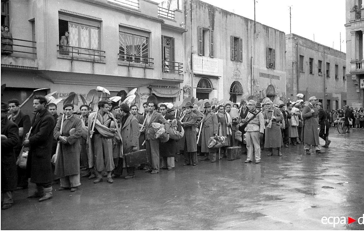 الصورة تظهر مجموعة من الأشخاص في صف طويل، يبدو أنهم ينتظرون شيئًا ما. يرتدون ملابس شتوية، ويحملون حقائب وأشياء أخرى. في الخلفية، يمكن رؤية مبانٍ تاريخية بشرفات ونوافذ. الجو يبدو هادئًا، ولكن المحيط يوحي بوجود حدث مهم أو تجمع. قد تعكس الصورة فترة زمنية معينة أو حدث اجتماعي.