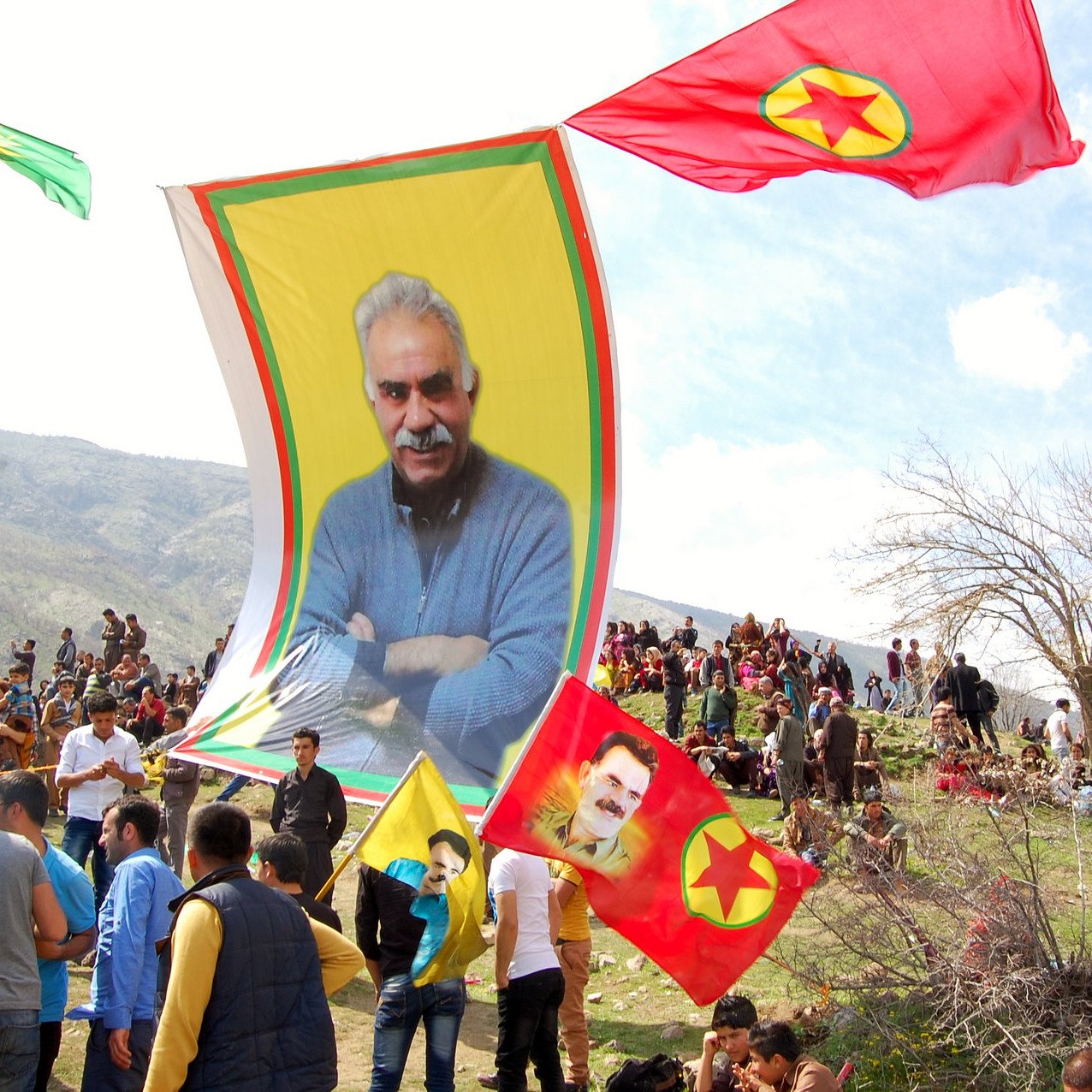 L'image montre une grande réunion en plein air, probablement lors d'une célébration ou d'un événement politique. On peut voir un grand drapeau avec le portrait d'un homme, entouré de plusieurs drapeaux aux couleurs vives, notamment du jaune et du rouge. De nombreuses personnes sont rassemblées et semblent animées, certaines brandissant des drapeaux. En arrière-plan, des montagnes et des arbres se dressent, suggérant un cadre naturel. L'atmosphère semble festive et engagée.