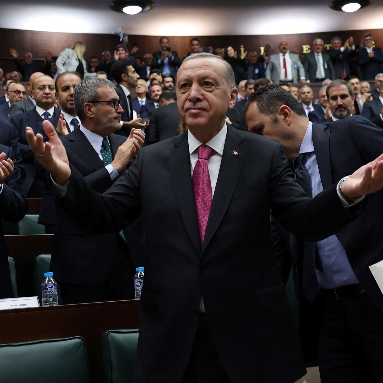 L'image montre un homme en costume, les bras écartés, se tenant au centre d'une assemblée constituée de nombreuses personnes applaudissant autour de lui. Il semble apprécier l'attention du public, dans un environnement de type parlementaire, où les membres sont assis sur des bancs et affichent des expressions variées. L'atmosphère est dynamique, suggérant un moment de célébration ou de discours important.