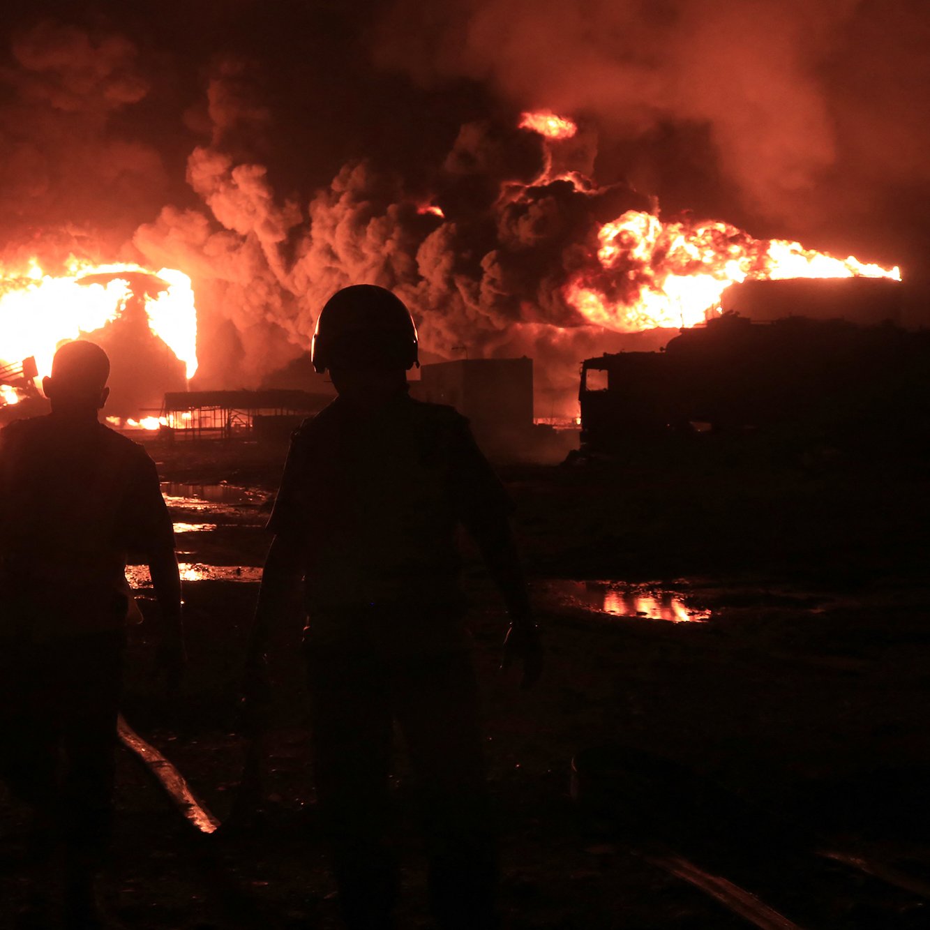 The image depicts two figures walking away from a large fire at night, with bright flames and thick smoke rising into the sky. The scene conveys a sense of urgency and danger.