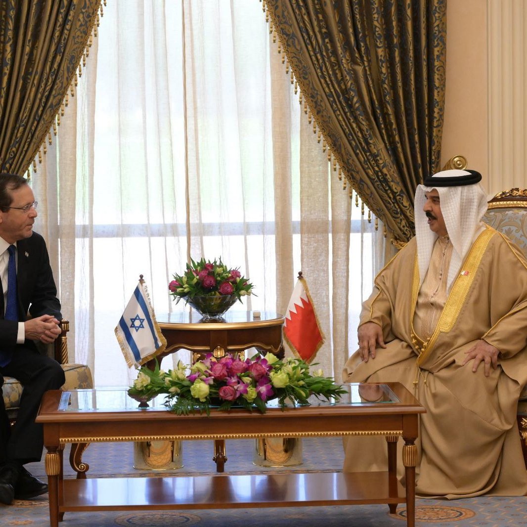Deux hommes en costume discutent assis dans un salon élégant, drapeaux visibles.