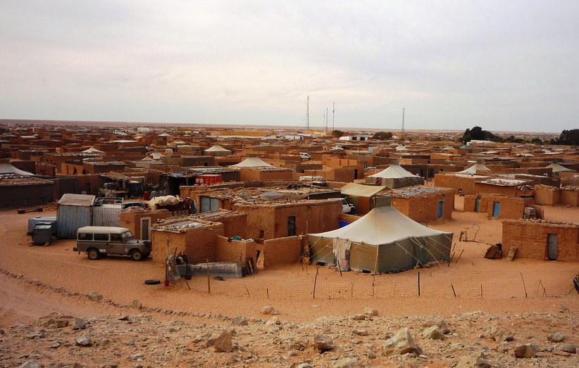 L'image présente un paysage désertique où l'on aperçoit un village de tentes et de maisons en argile. Les structures sont éparpillées dans le sable, entourées d'un terrain aride. Au fond, quelques antennes sont visibles, suggérant une connexion à l'extérieur. Le ciel est nuageux, créant une ambiance calme et légèrement mélancolique.