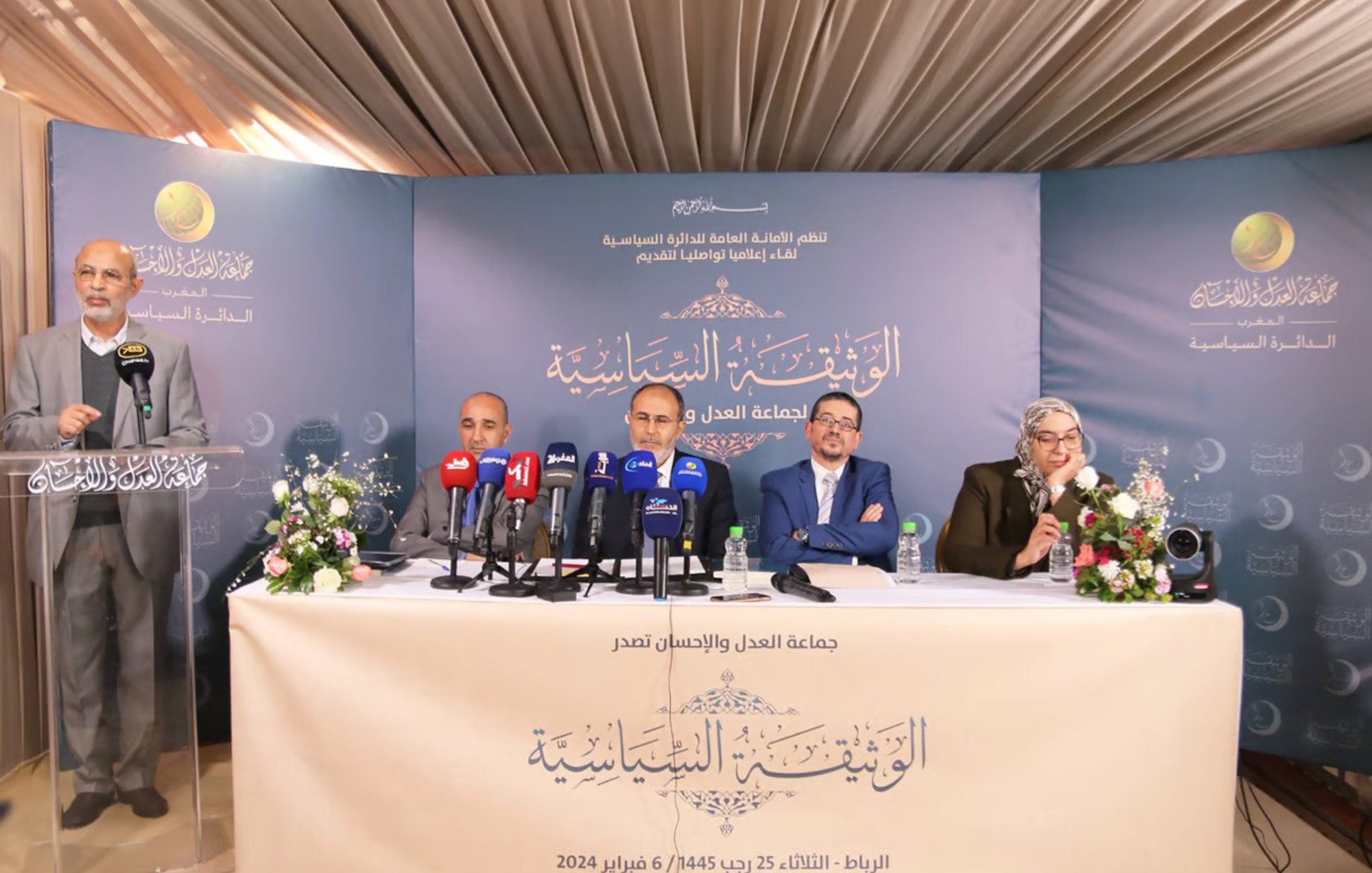 L'image montre une conférence de presse avec plusieurs intervenants assis derrière un pupitre. Ils sont entourés de micros et de caméras, indiquant une attention médiatique. Des fleurs et une décoration soignée ajoutent une touche de professionnalisme à l'événement. On peut voir un intervenant debout, probablement en train de parler ou de présenter, tandis que les autres écoutent attentivement. Le fond de la scène est orné de motifs ou de logos, suggérant que c'est un événement officiel ou formel.