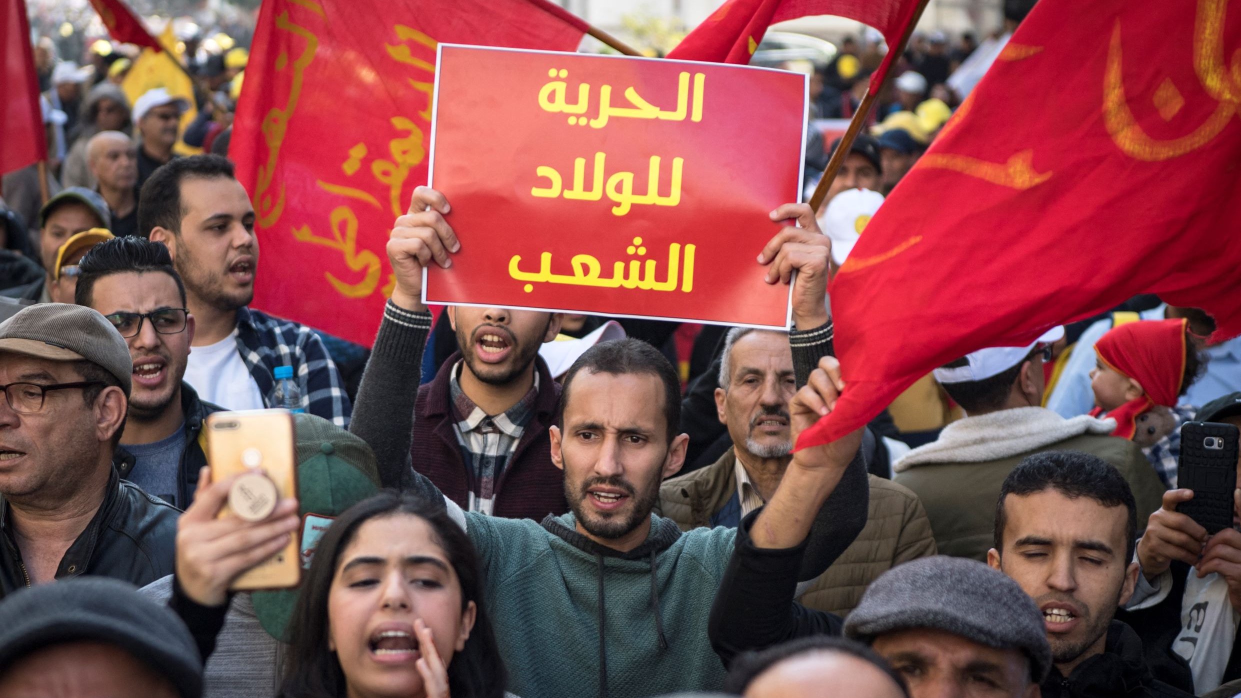 تظهر الصورة مجموعة من الأشخاص في مسيرة احتجاجية. يحمل أحد المتظاهرين لافتة مكتوب عليها "الحرية للولد الشعب"، بينما يبدو أن الحشود من حوله ترفع أعلامًا حمراء. التعبيرات في وجوه المتظاهرين تتراوح بين الحماس والعزيمة، مما يدل على روح التضامن والمشاركة في المطالبة بالحقوق. الجو العام يظهر فيه نشاط وحيوية، مما يشير إلى أهمية الرسالة التي يسعون لنقلها.