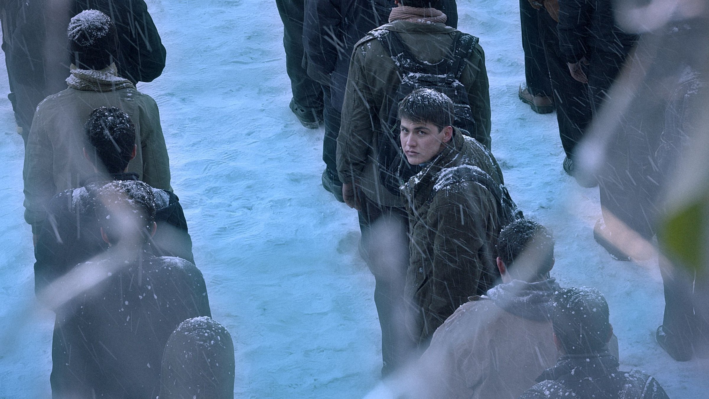 L'image montre une scène froide et enneigée avec une foule de personnes, des flocons de neige tombant. Au centre, un jeune homme se retourne, son visage est légèrement visible. Les autres autour de lui sont flous et vêtus de manteaux sombres, créant une atmosphère de tension et d'incertitude.