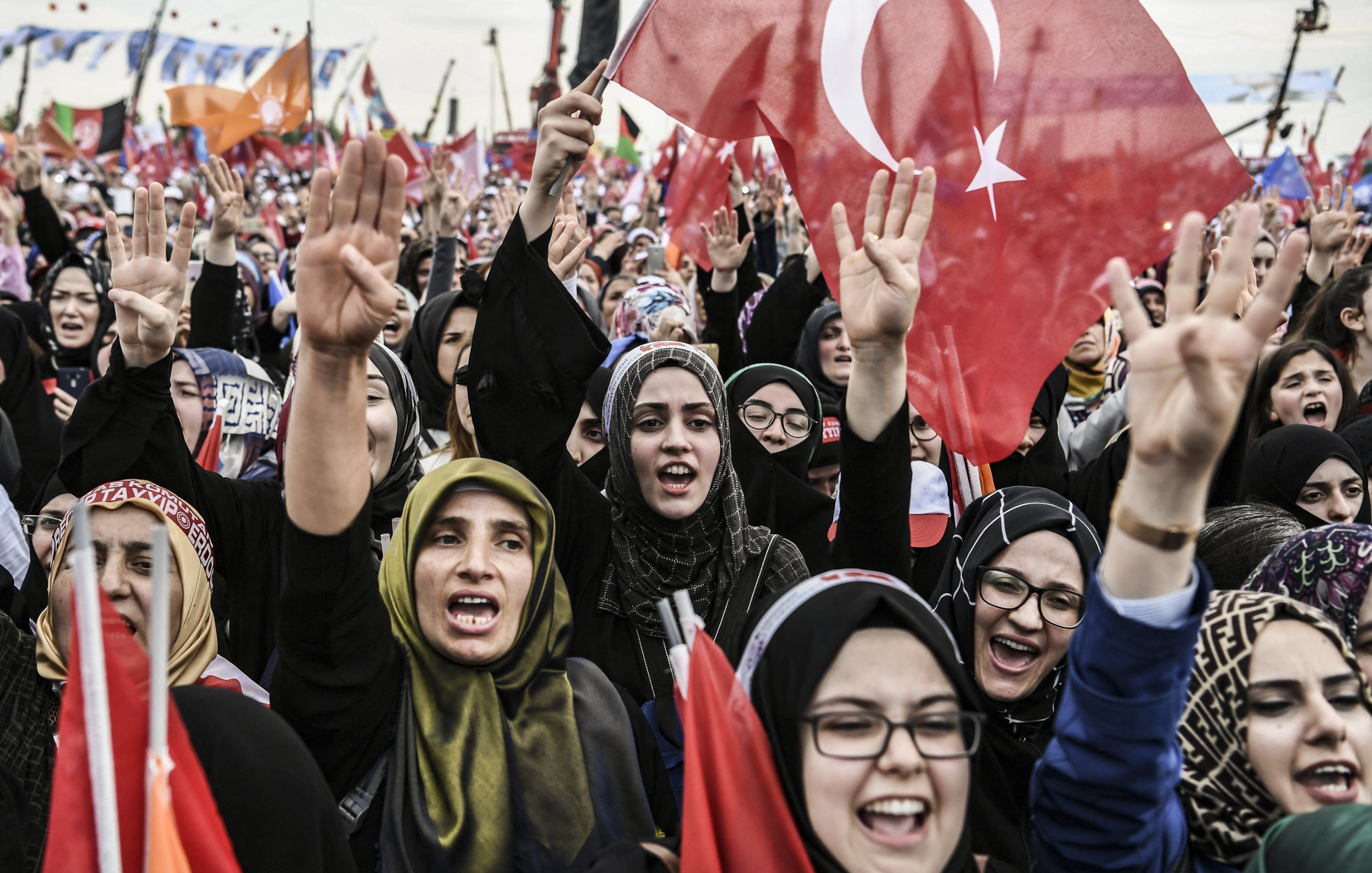 L'image montre une grande foule de personnes, principalement des femmes, qui sont rassemblées lors d'un événement ou d'une manifestation. Elles expriment leur enthousiasme et leur soutien en levant des drapeaux, dont certains arborent le symbole de la Turquie. Les participantes portent des vêtements traditionnels ou des foulards, et certaines semblent chanter ou crier des slogans. L'ambiance est dynamique et remplie d'énergie collective, suggérant une atmosphère de célébration ou de revendication.