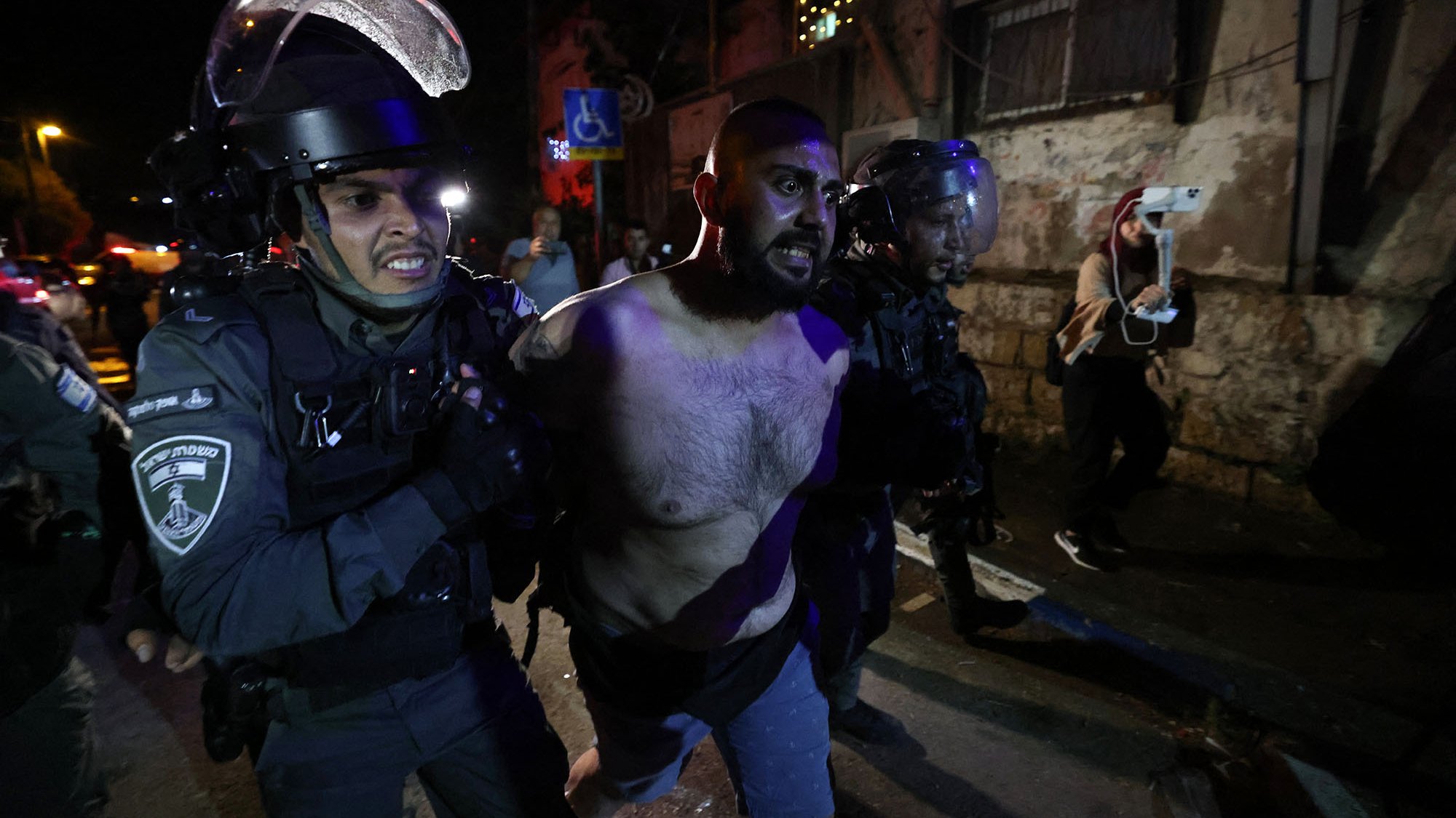 L'image montre des policiers en uniforme, portant des casques et des protections, en train de maîtriser un homme. L'homme est torse nu et semble être en train d'être arrêté. L'environnement nocturne, avec des lumières et des bâtiments en arrière-plan, suggère une situation de tension, potentiellement liée à des manifestations ou des troubles civils. Les expressions et la posture des policiers indiquent une approche ferme envers la situation.
