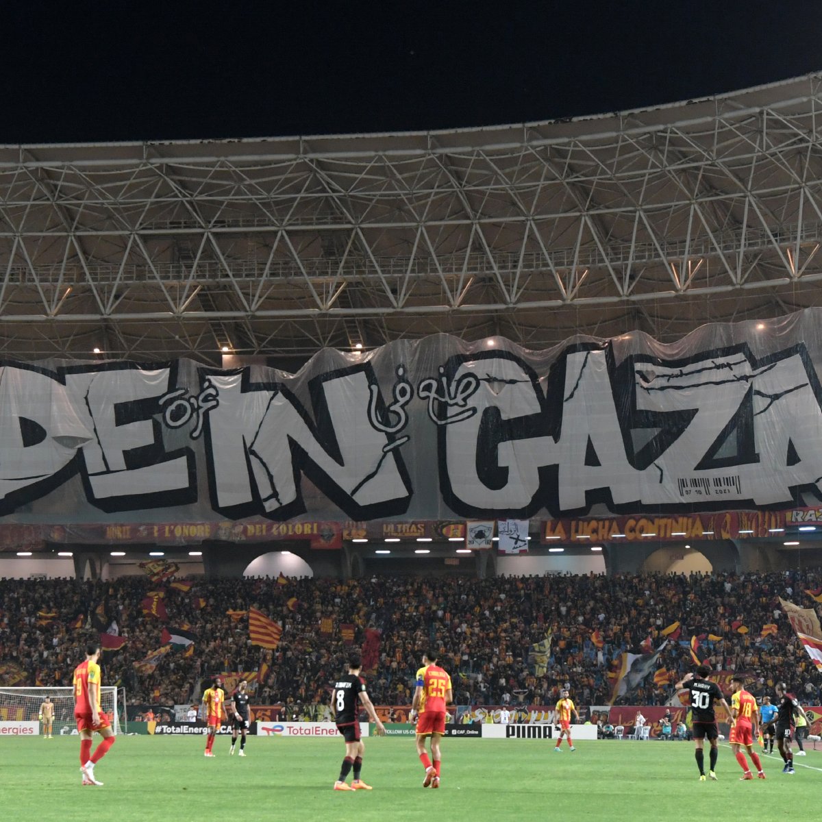 L'image montre un stade de football avec une grande banderole affichée. La banderole est ornée du texte "MADE IN GAZA" écrit en lettres stylisées et imposantes. Le stade est rempli de supporters, et l'atmosphère semble animée. On peut voir des joueurs sur le terrain, ce qui indique qu'un match est en cours. L'ambiance est festive et engagée.