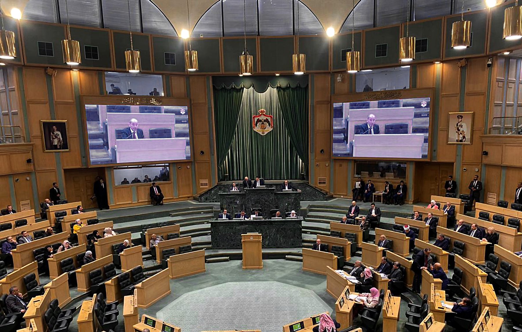 The image depicts an interior view of a legislative assembly or parliament. The chamber is spacious, featuring a semi-circular arrangement of desks for members. At the front, there is a podium where a speaker or official is addressing the assembly. Large screens on either side display information or the speaker to the attendees. The walls are adorned with drapes and emblems, contributing to a formal atmosphere. Members of the assembly are seated, engaged in discussion or listening attentively. The overall setting is one of a governmental deliberation space, likely during a session or meeting.