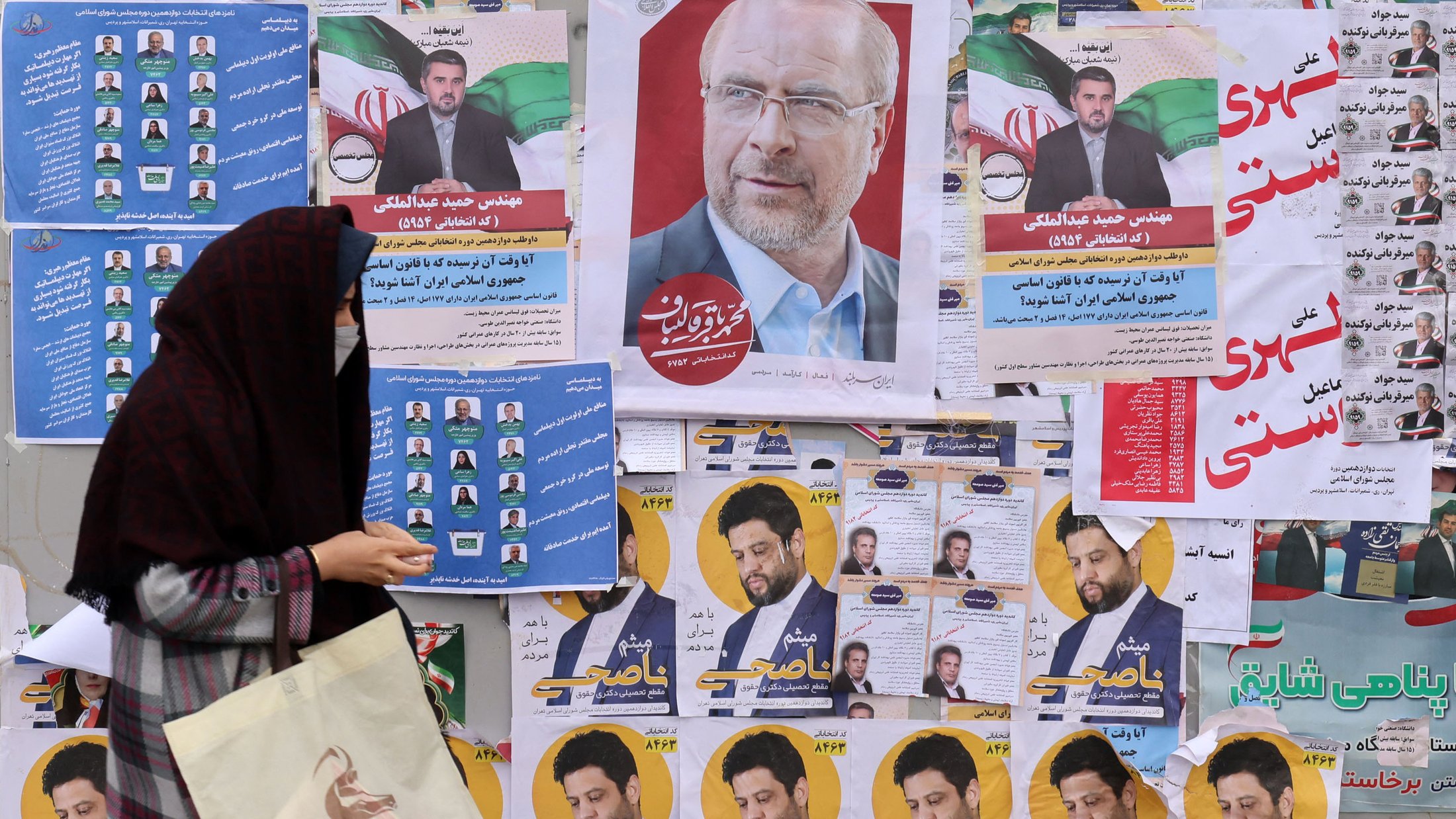 L'image montre un mur recouvert de diverses affiches électorales. On peut voir plusieurs portraits de candidats, accompagnés de slogans et d'informations. Au premier plan, une femme portant un hijab et tenant un sac semble marcher devant ces affiches, ce qui suggère une ambiance électorale. Les couleurs des affiches sont vives et variées, ajoutant à l'aspect animé de la scène.