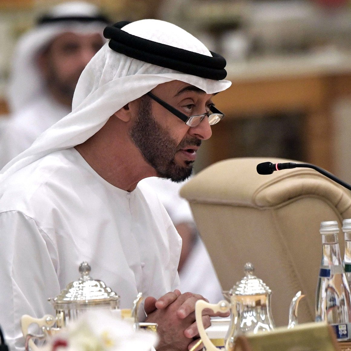 L'image montre un homme portant une tenue traditionnelle émiratie, comprenant une kandura blanche et un ghutrah. Il est assis à une table, apparemment en train de s'adresser à un public. À côté de lui, on peut apercevoir des carafes et des verres d'eau. Le décor semble être celui d'une réunion ou d'une conférence, avec d'autres personnes en arrière-plan. L'atmosphère est sérieuse et formelle.